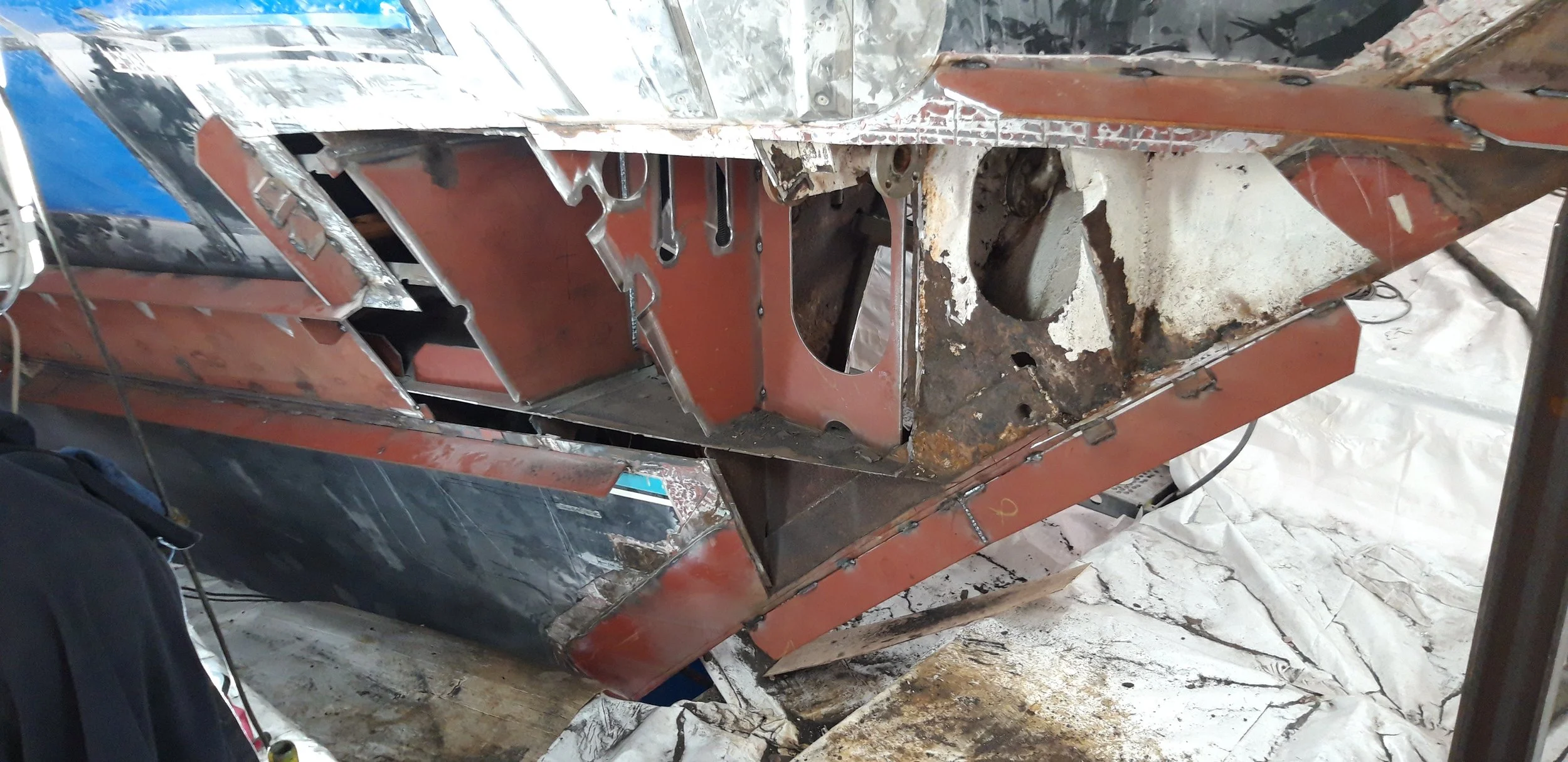 Close-up view of a car body undergoing restoration, showing rusted sections, exposed metal, and new metal panels being fitted.