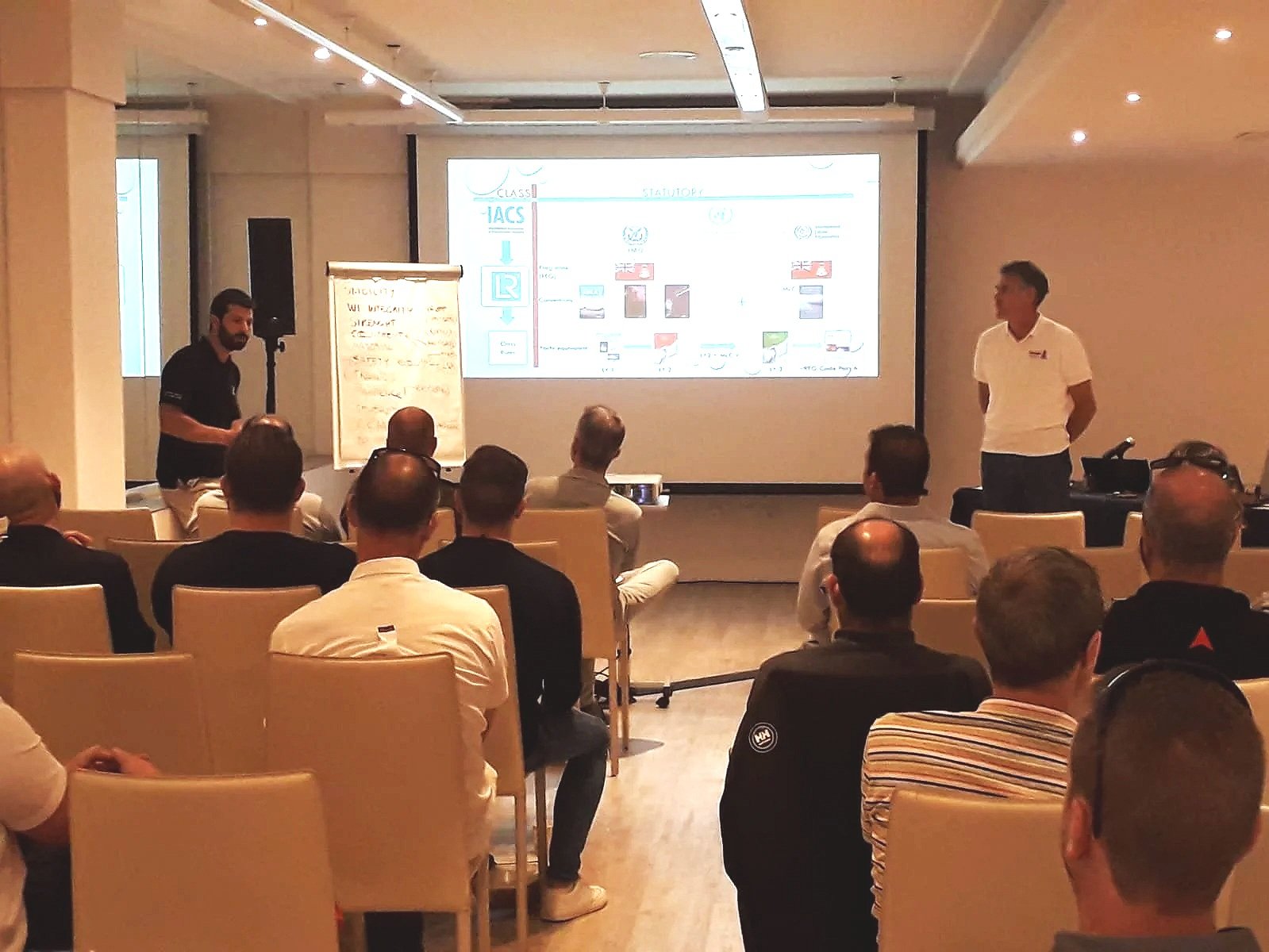A group of people attending a presentation in a conference room. Two men stand at the front, one near a large screen with a presentation slide and the other near a whiteboard with notes. The audience is seated in rows facing the presenters.