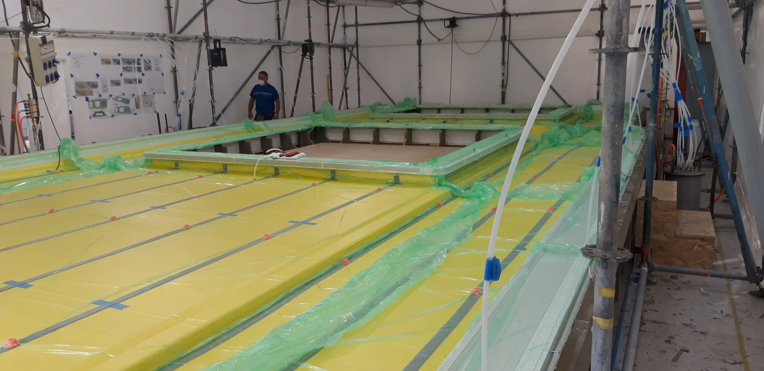 Interior construction site with a large, rectangular foam core structure outlined by green tape and plastic sheeting, surrounded by scaffolding; a worker in a blue shirt and face mask stands nearby.