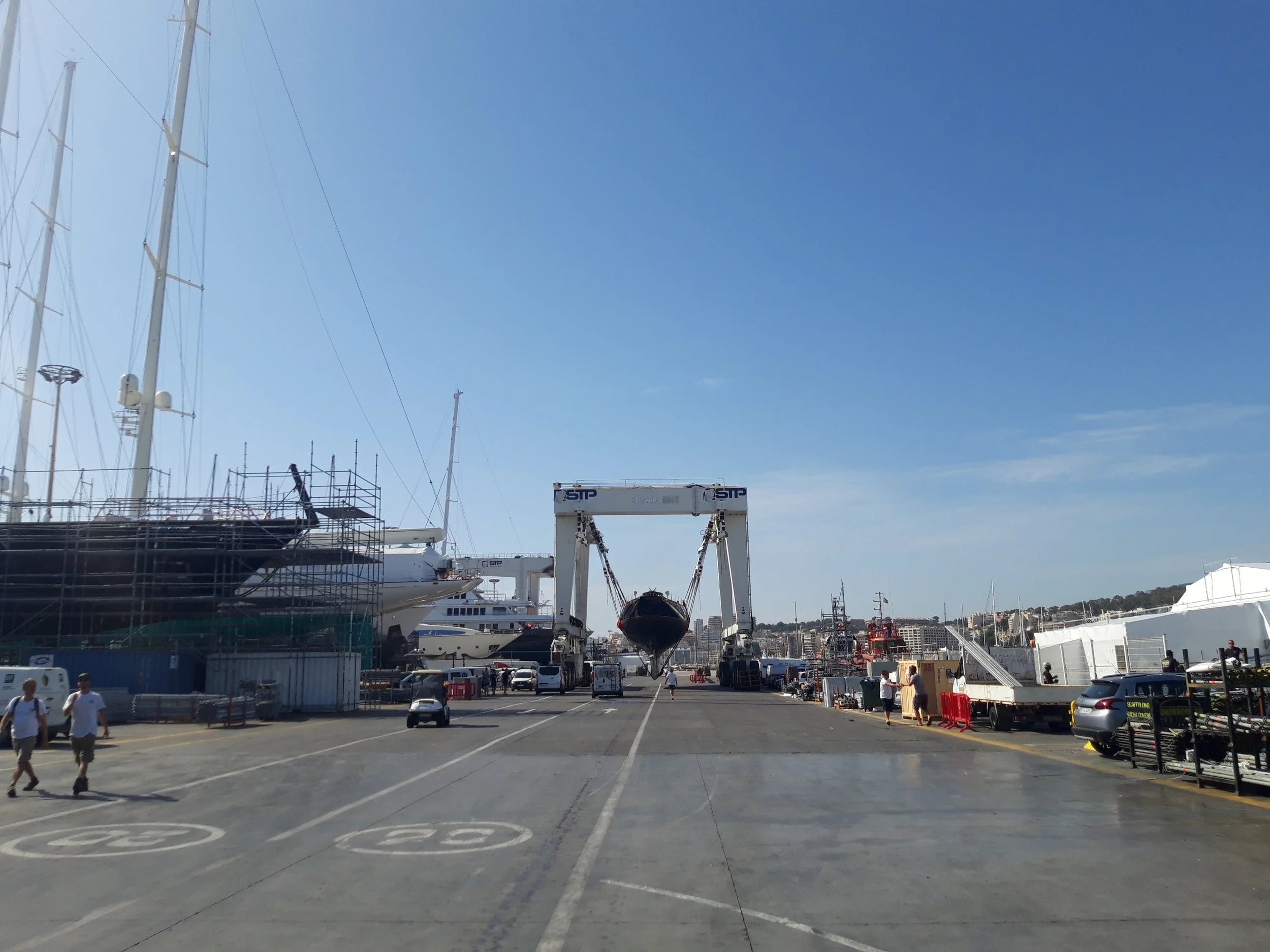 A boat being lifted out of the water by a large crane at a marina with yachts docked on either side, and a clear blue sky overhead.