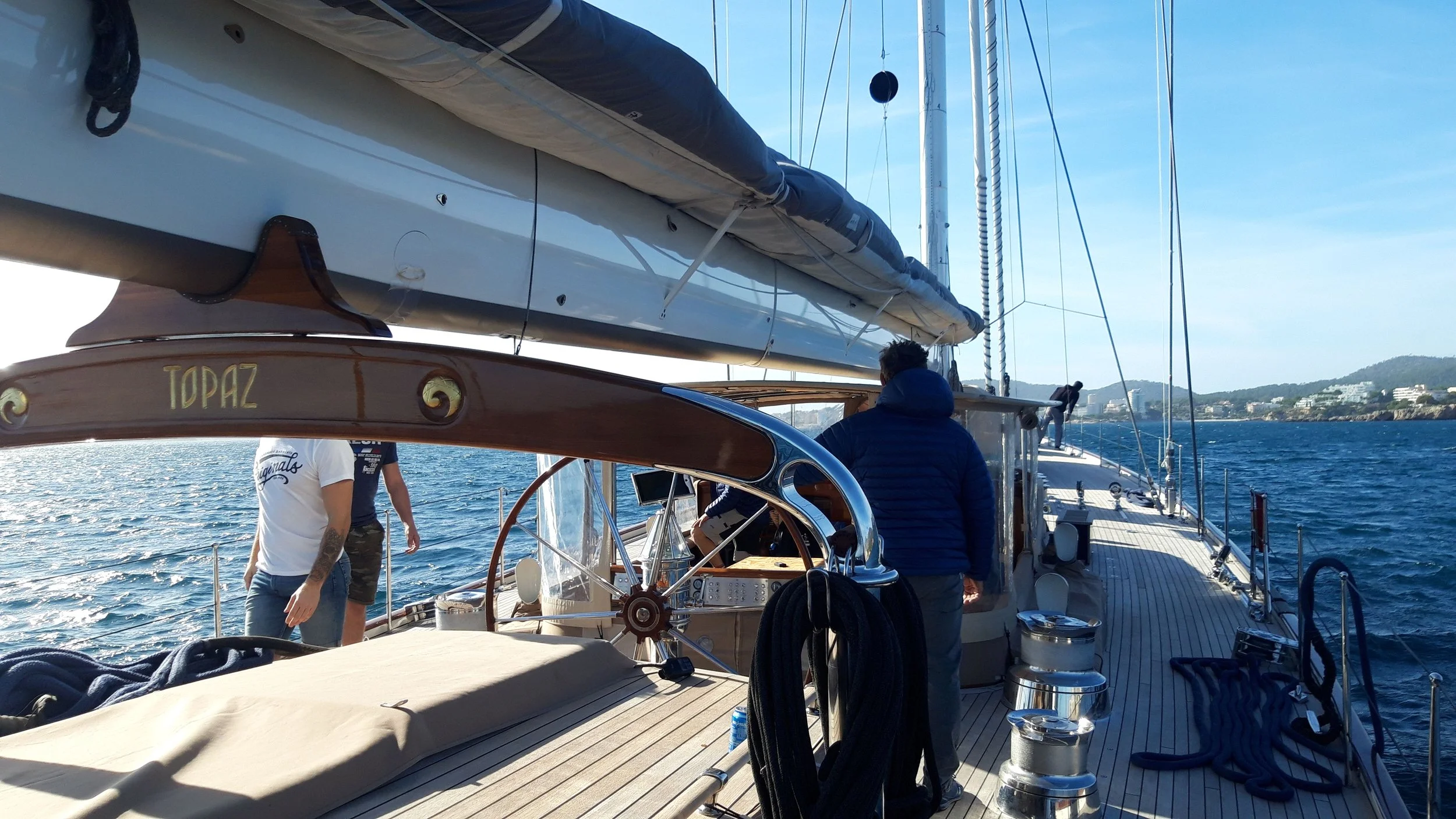View of a sailboat navigating on a body of water with a clear sky and distant shoreline, showing the deck, steering wheel, sailors, and the boat's mast and rigging.