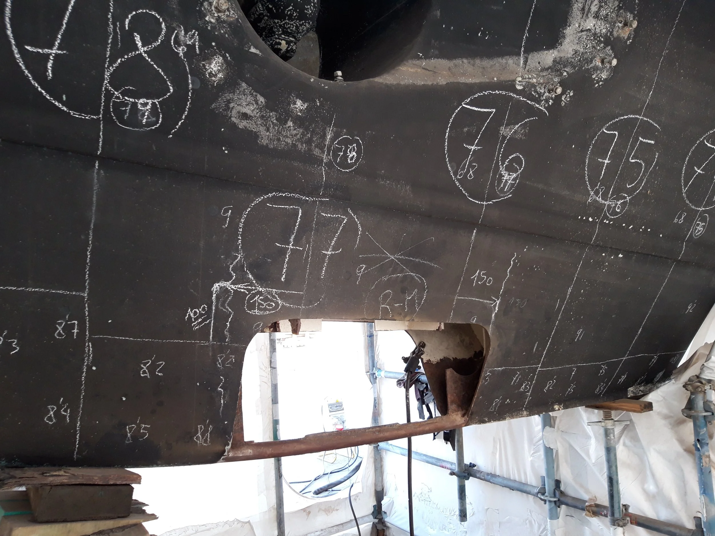 Numerical markings and drawings on a black metal surface, likely part of a ship or industrial equipment, with scaffolding and tools visible below and around.