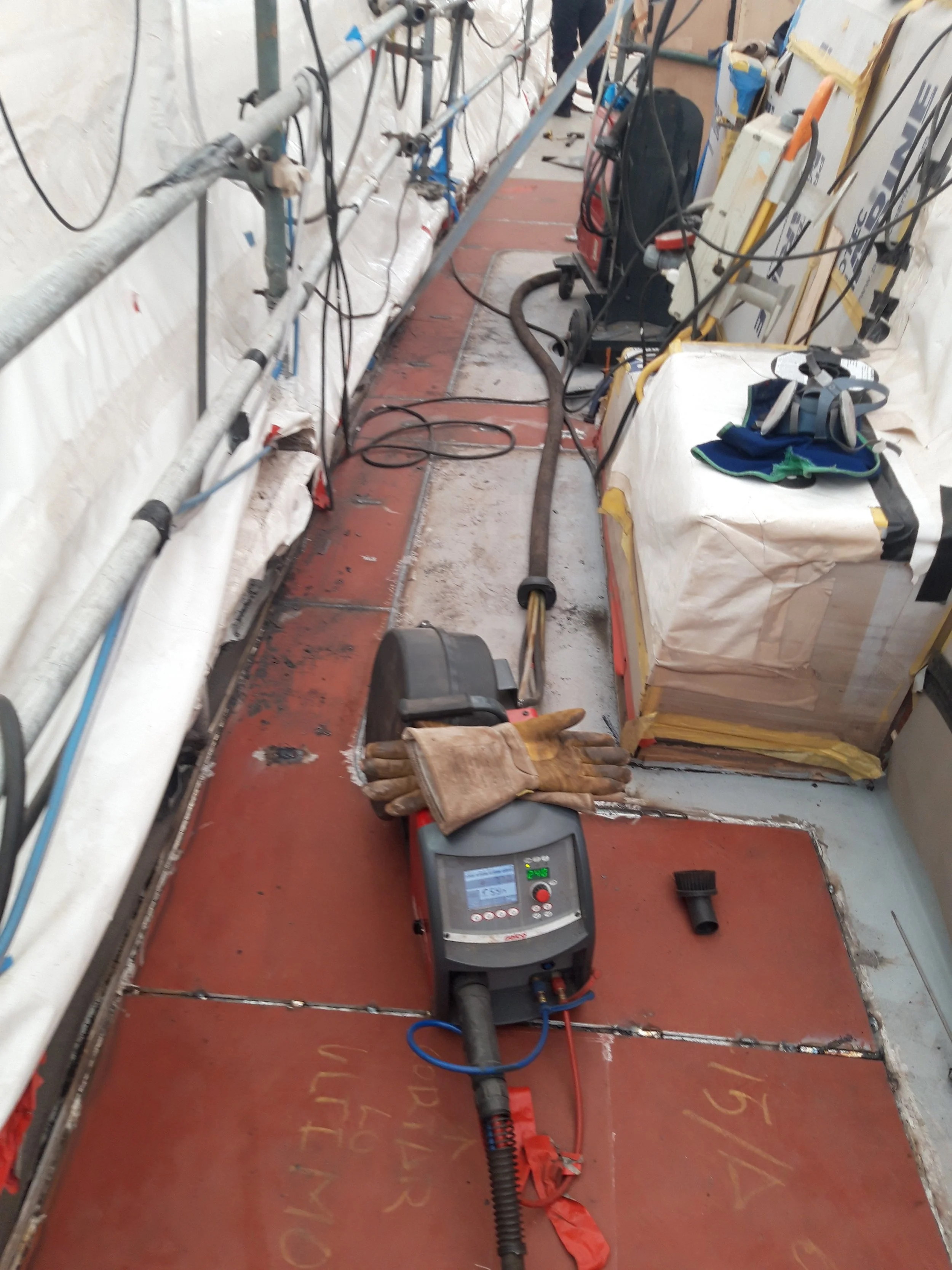Worksite with tools, equipment, and protective gear on the floor, including a hose, gloves, and a digital device, with construction materials and wiring along the walls.