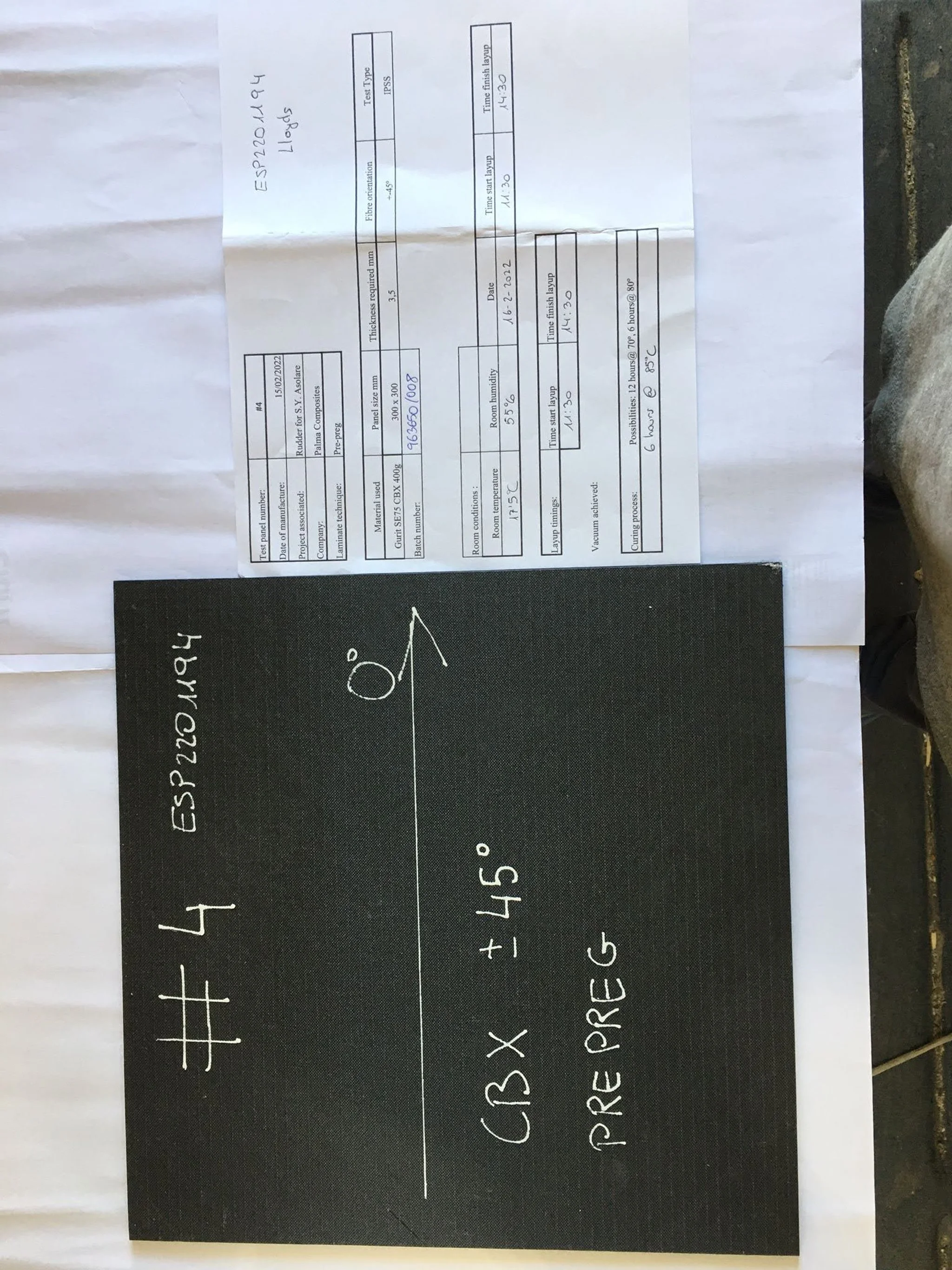 A blackboard with white chalk writing showing a formula: #4 (CRB X + 45° PRE G). Above the blackboard, there are two sheets of paper with technical details related to a project or product, including specifications, date, batch number, and other techn