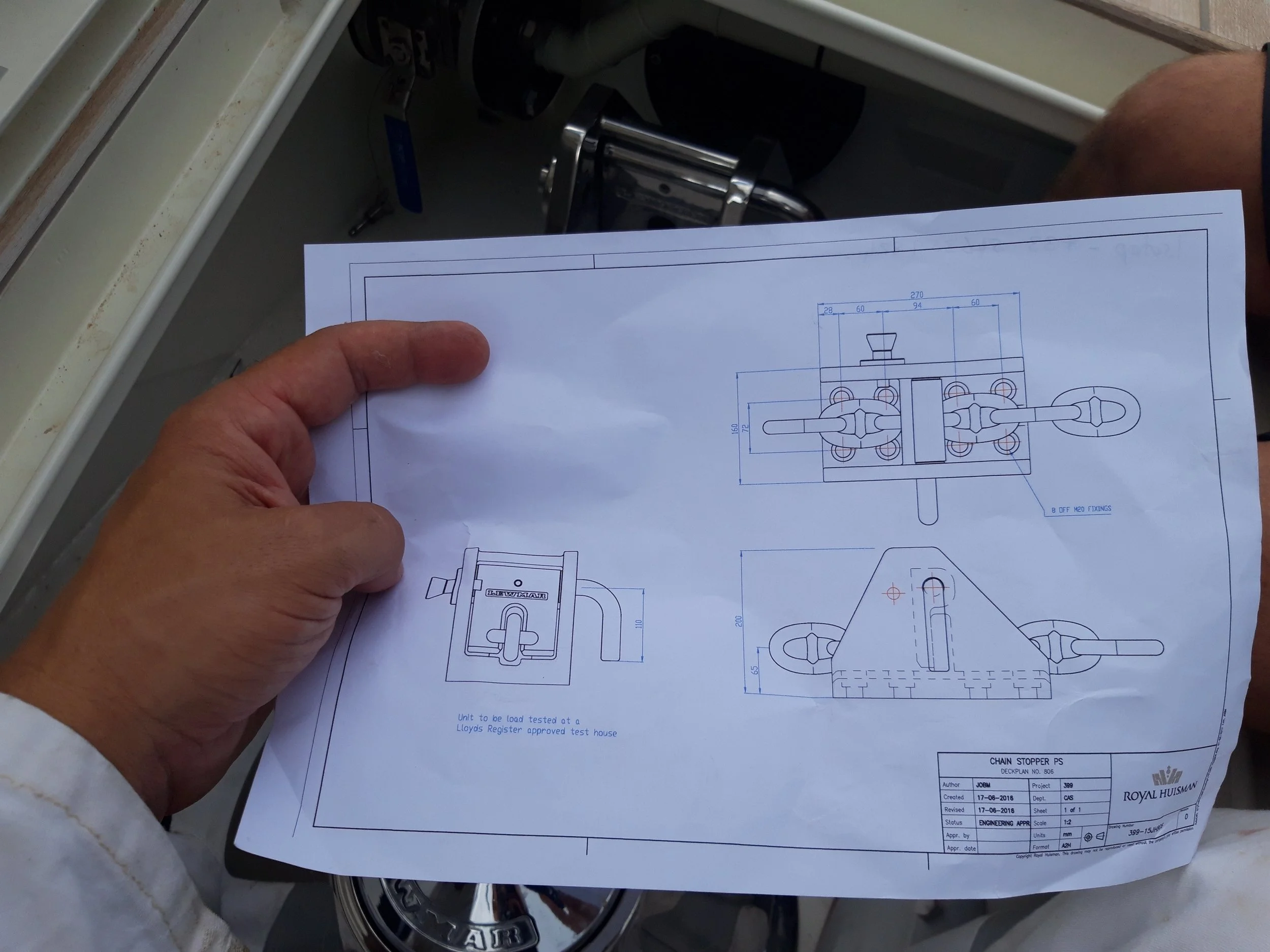 A person holding engineering blueprints for a chain stopper device, with technical drawings and measurements, over a work surface.