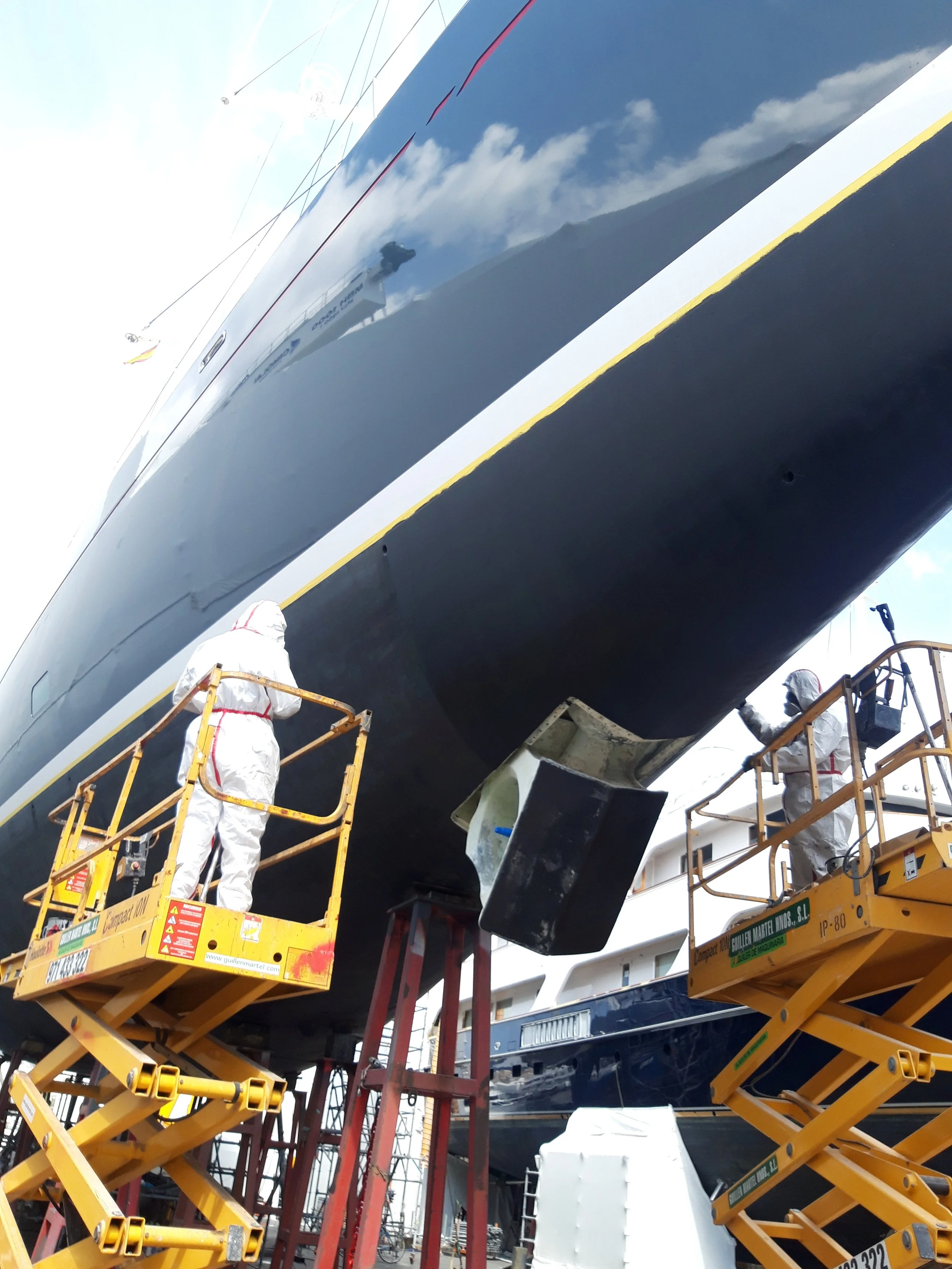 Workers in protective gear on lifts repairing or inspecting the hull of a large yacht, with a cloudy sky reflected on the shiny surface of the boat.