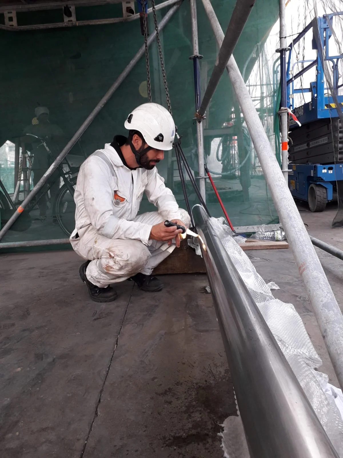 A worker in a white jumpsuit and safety helmet welding or grinding a metal pipe on a boat or ship under a scaffolding.