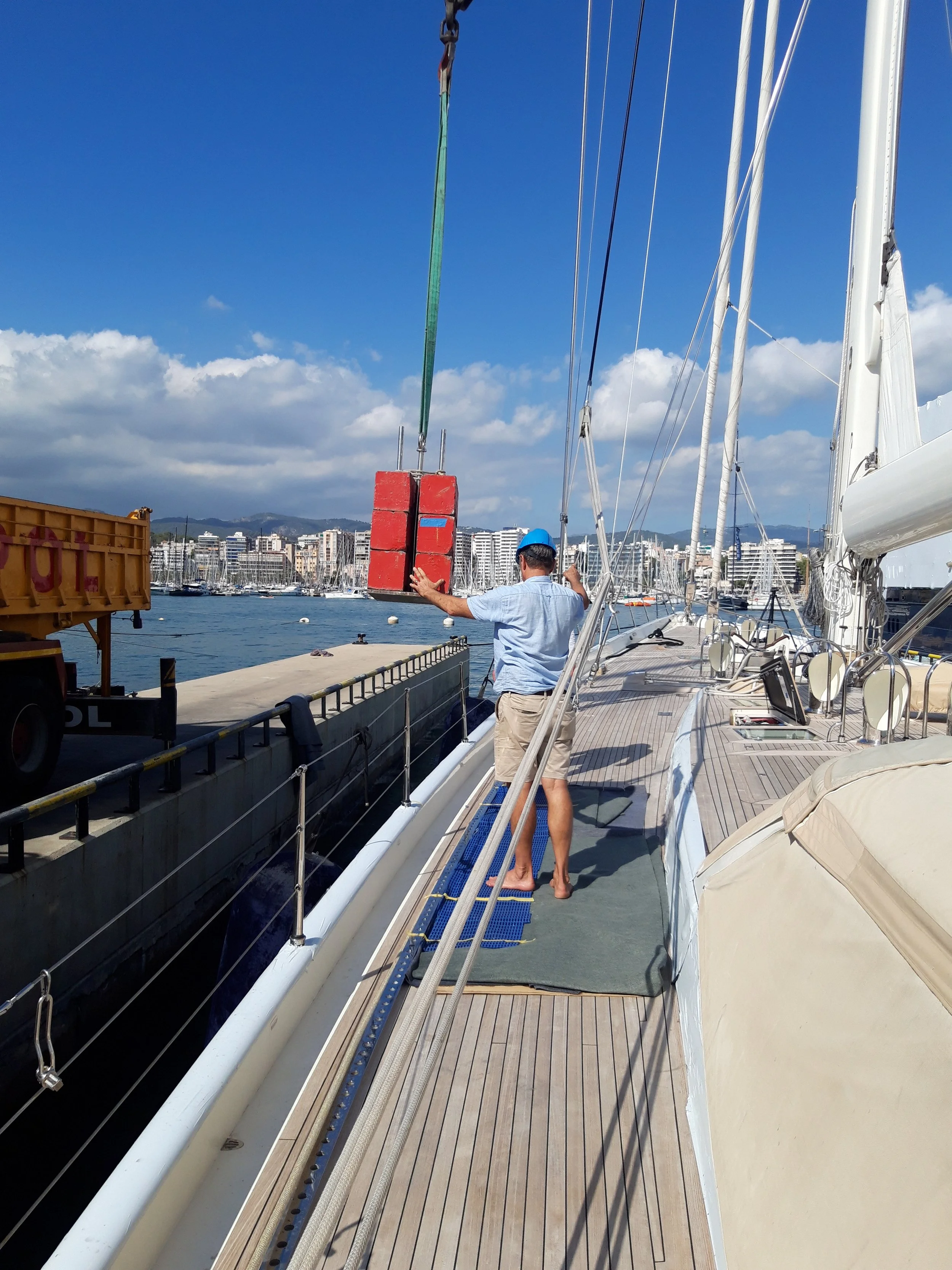 A man on a yacht yard, wearing a blue cap, light blue shirt, and khaki shorts, is handling a large red object in the boat's deck area with sailboats and a city skyline in the background.