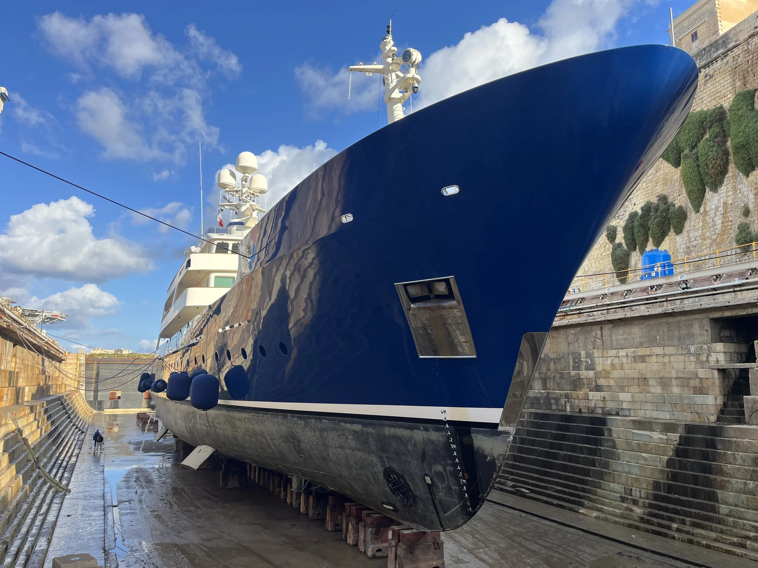 Large blue yacht in dry dock with a stone wall and sky in the background.