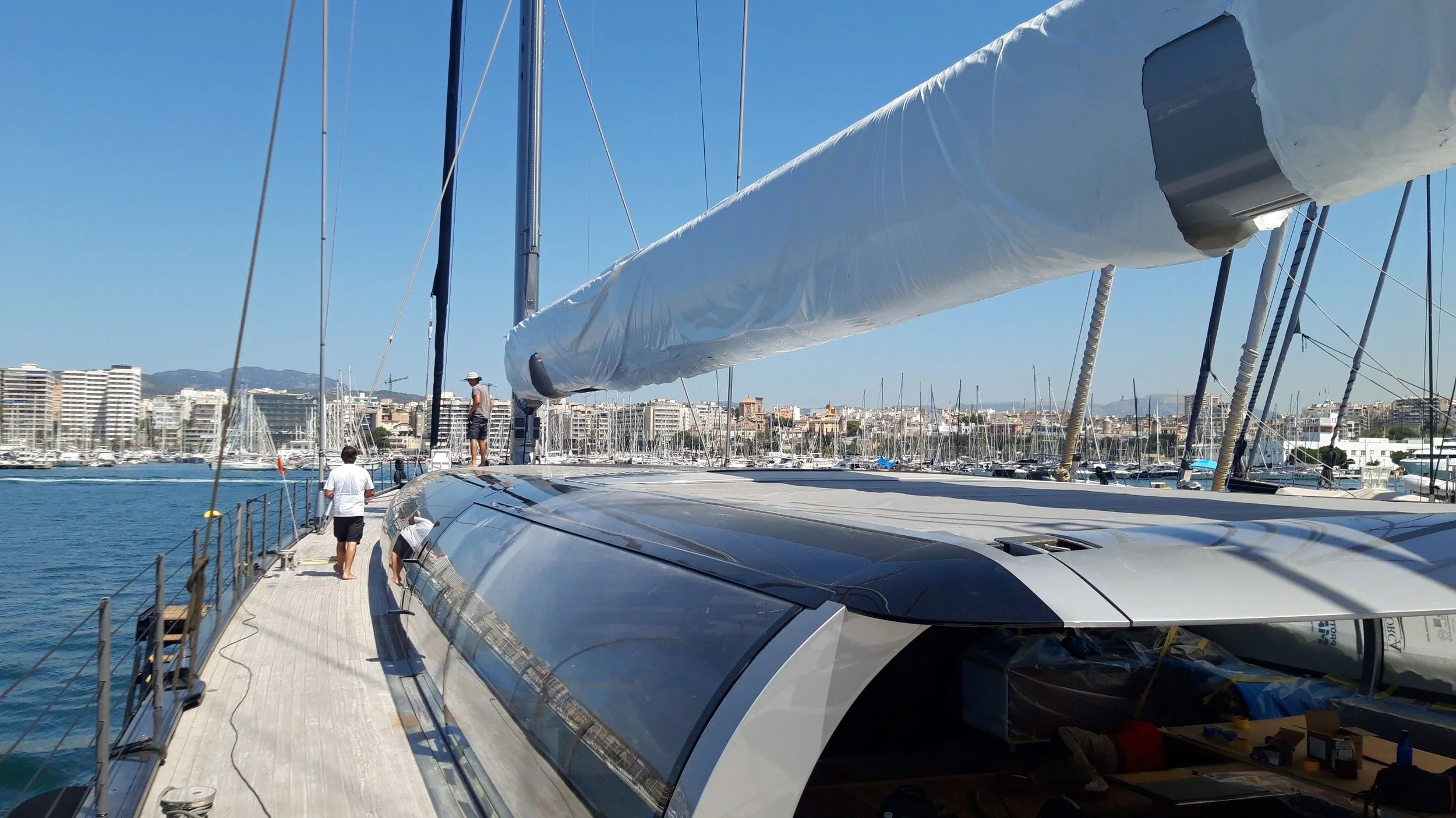 View of a modern sailboat docked at a marina with other boats and high-rise buildings in the background under a clear blue sky.