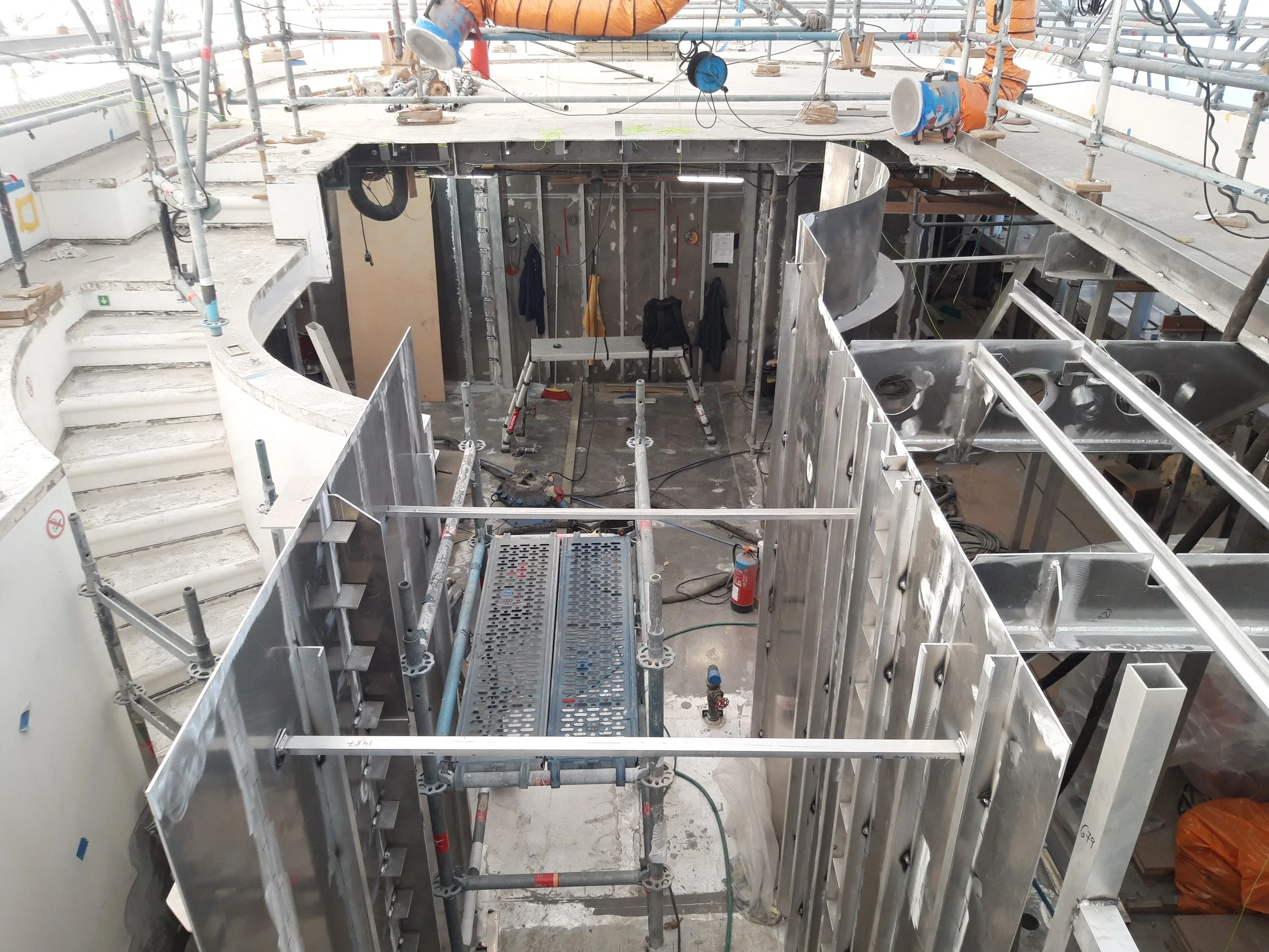 View of an indoor construction site, showing metal framework, scaffolding, and stairs with ongoing building work, including various construction tools and equipment.
