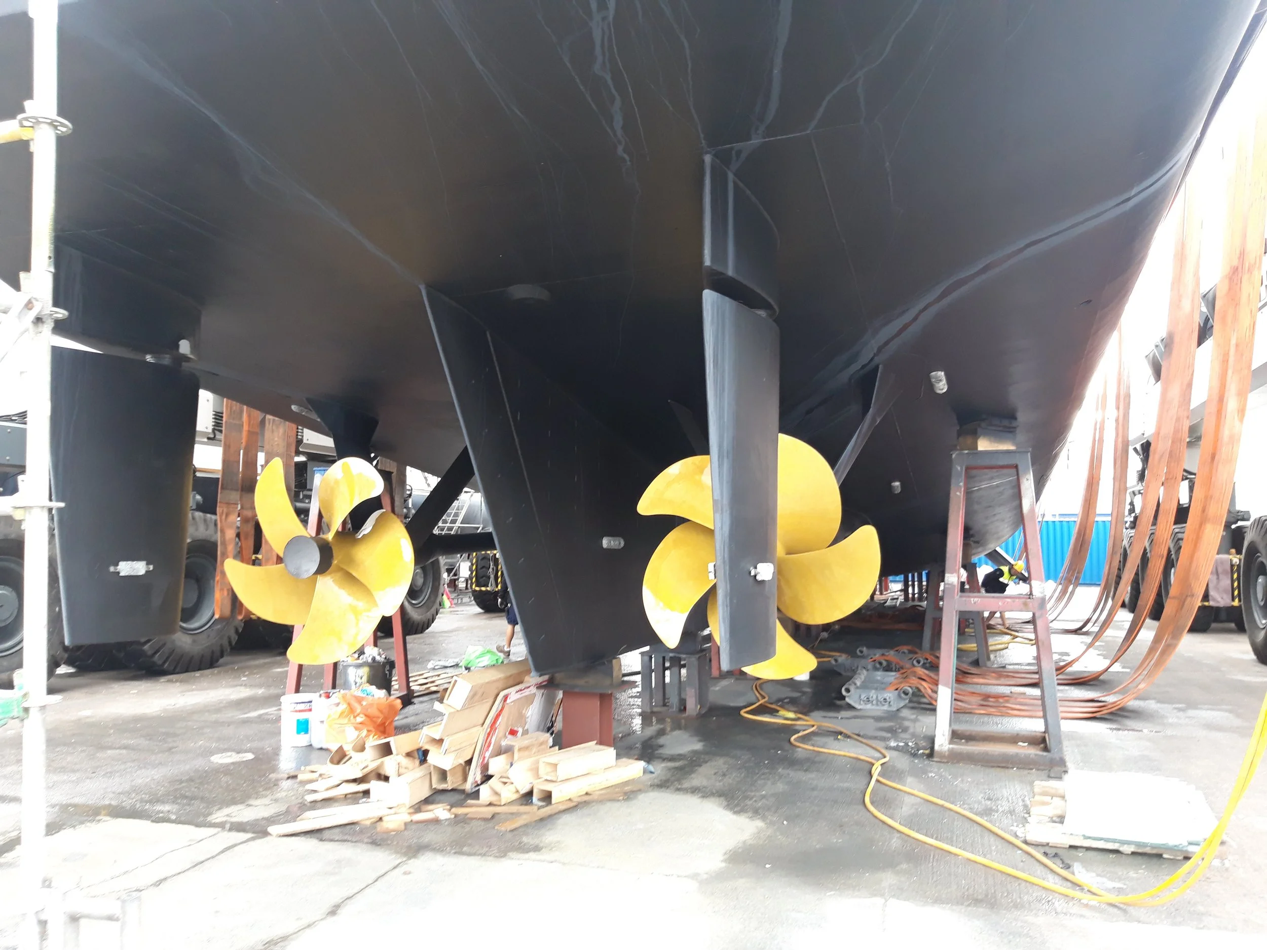 The underside of a large boat with two yellow propellers, surrounded by tools, ladders, and other equipment in a boatyard or dry dock.