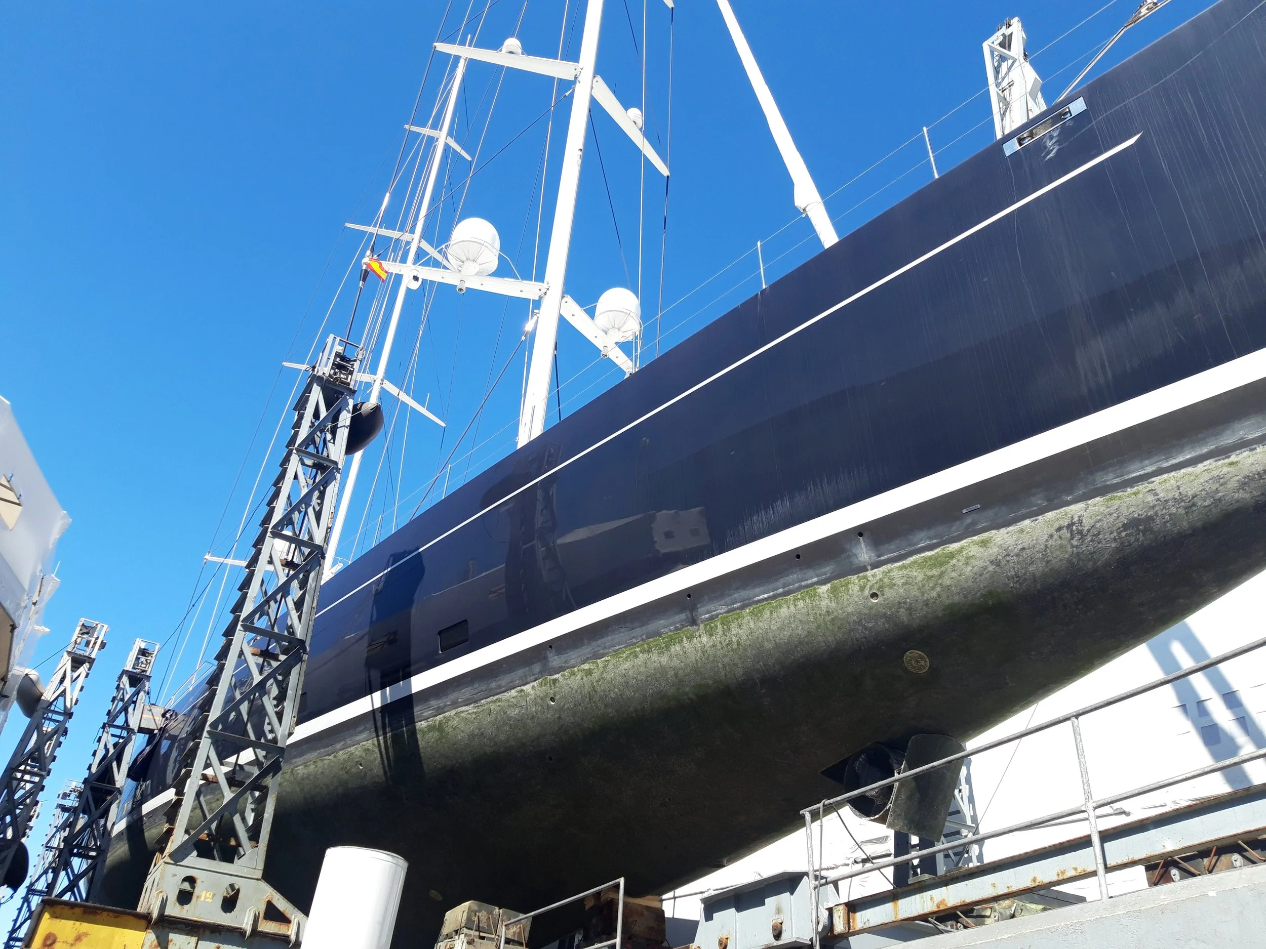 Large black sailboat on dry dock with scaffolding and maintenance equipment underneath, under a clear blue sky.