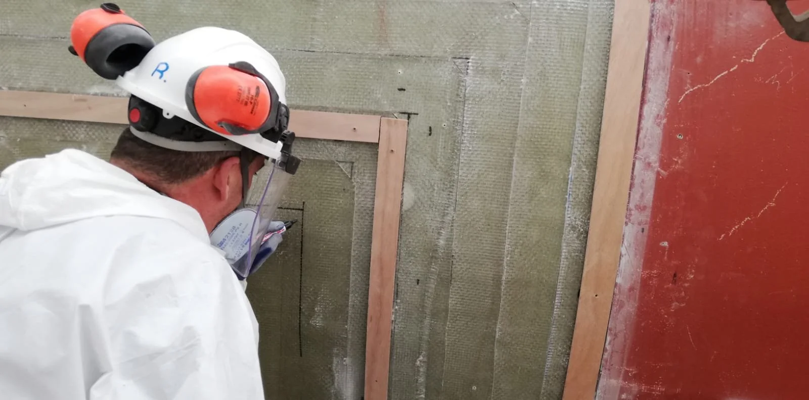 A construction worker wearing a white safety helmet, yellow ear protection, and a white protective suit is working on a wall, possibly installing or repairing drywall.
