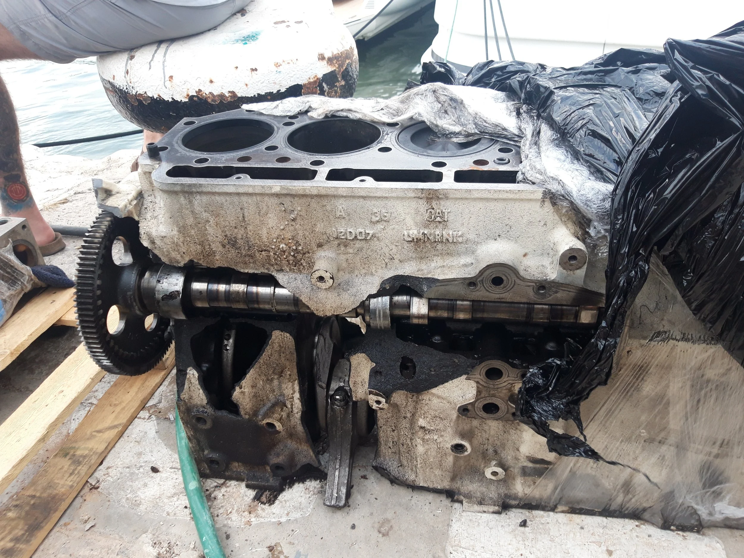 Outboard engine block resting on a wooden pallet, partially disassembled and dirty, with tools and boat parts nearby, at a dock.