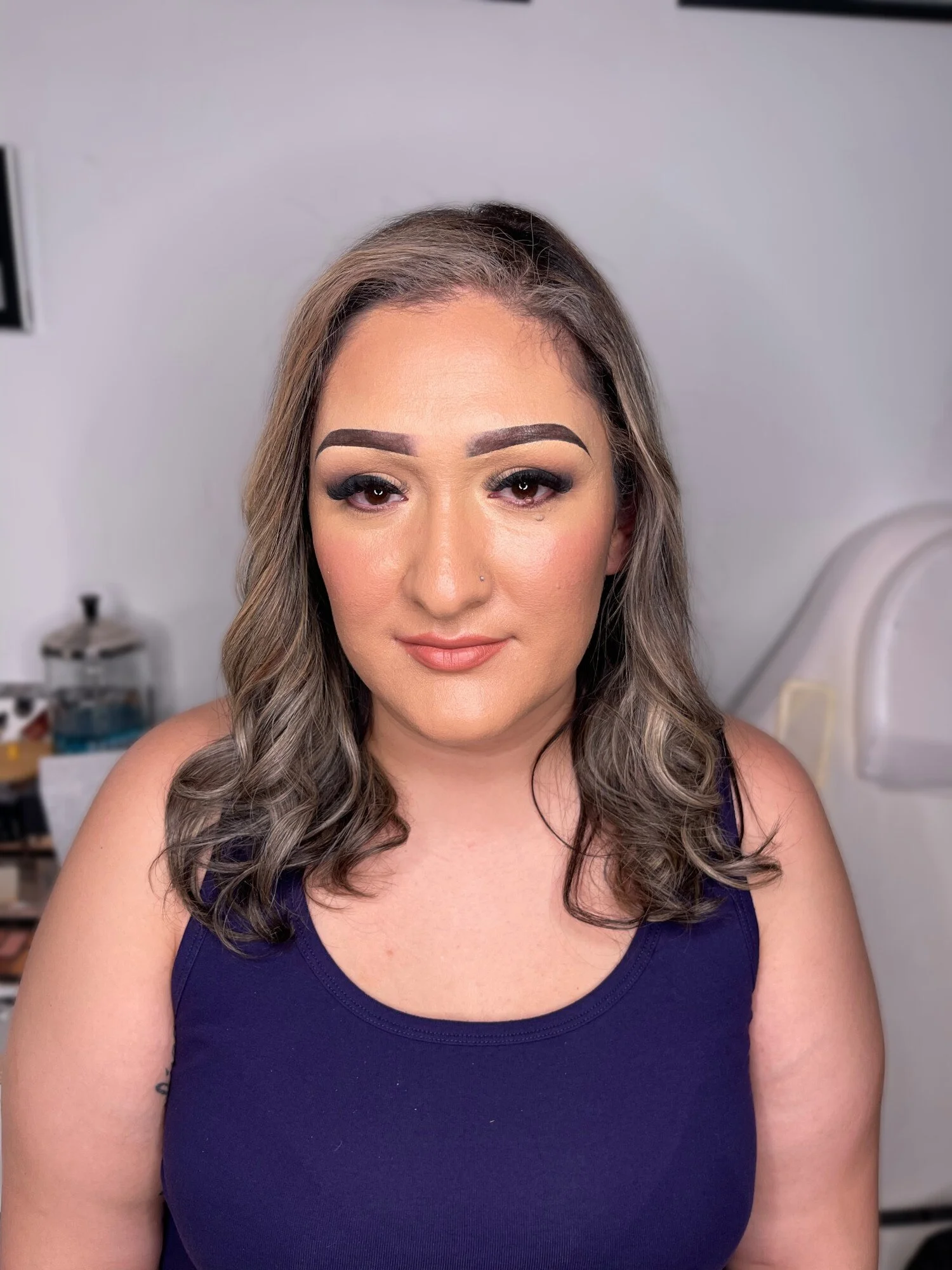 A woman with shoulder-length wavy hair and makeup, wearing a navy blue tank top, smiling softly in a room with a white wall background.