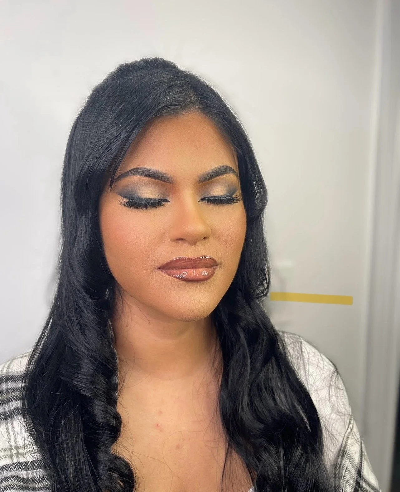 A woman with long black wavy hair and makeup with light eyeshadow, winged eyeliner, and glossy lipstick, with her eyes closed.