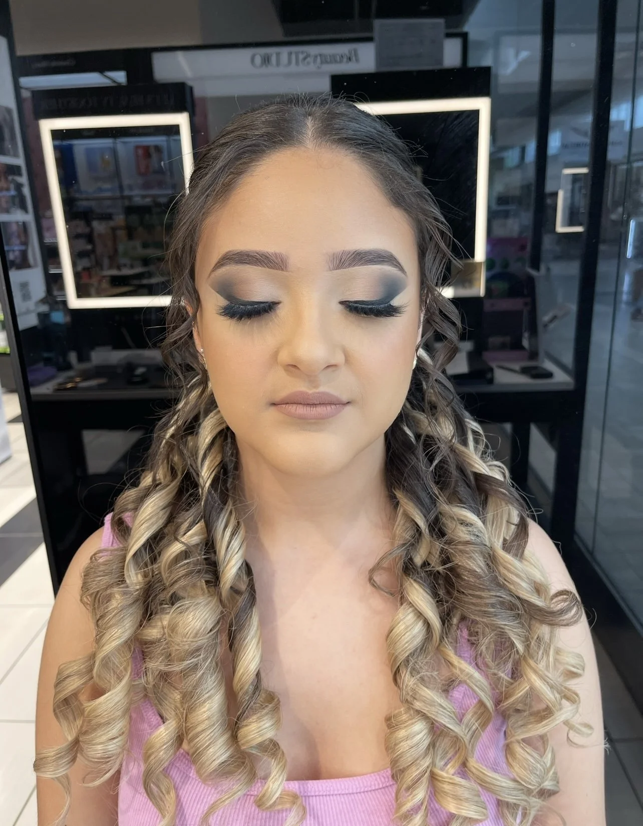 Woman with long blonde curled hair wearing makeup, closed eyes, in a makeup studio.