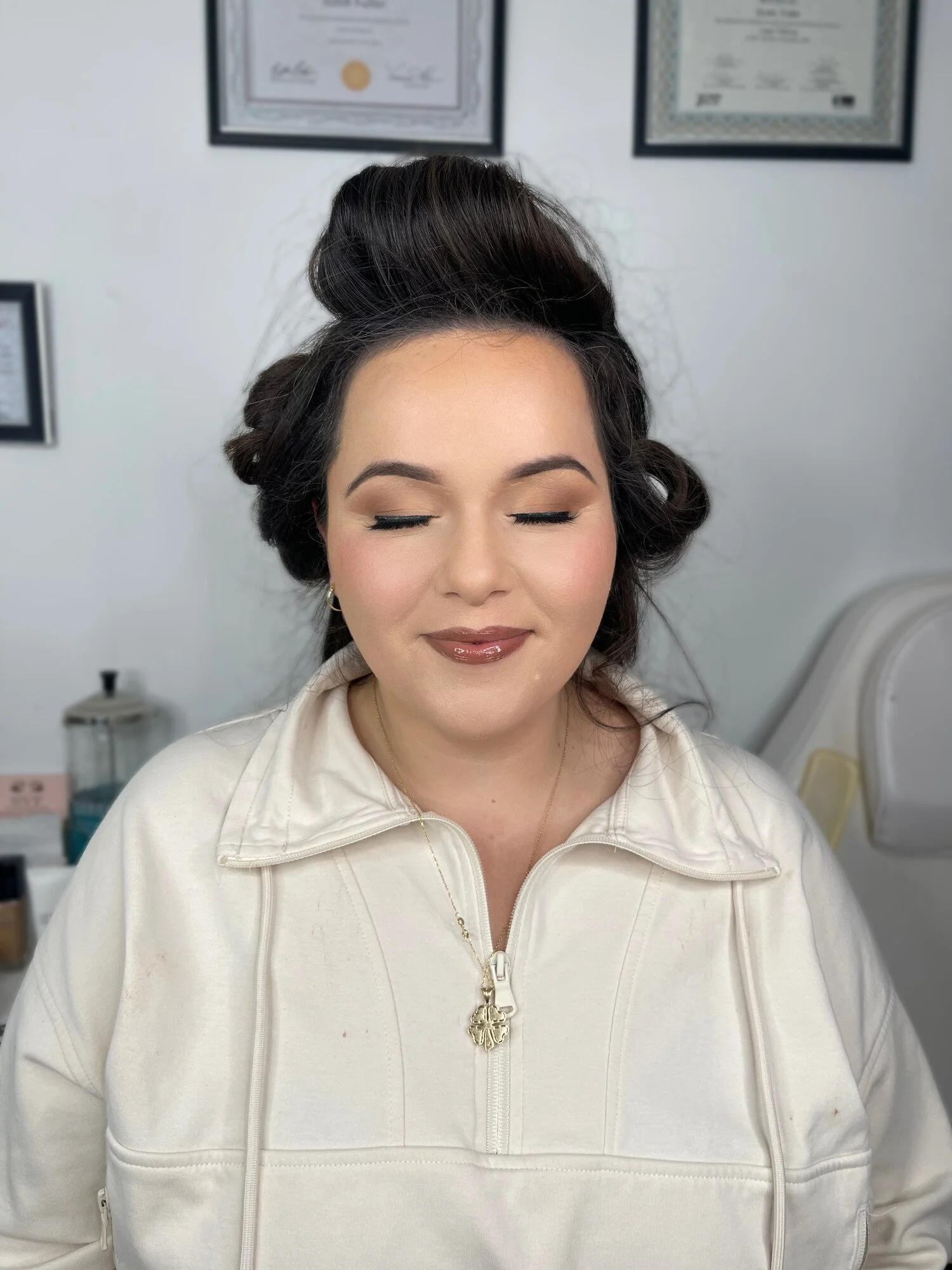 A woman with dark hair styled in vintage curls, wearing makeup, a white zip-up jacket, and a gold necklace, with her eyes closed, in a room with framed certificates on the wall.