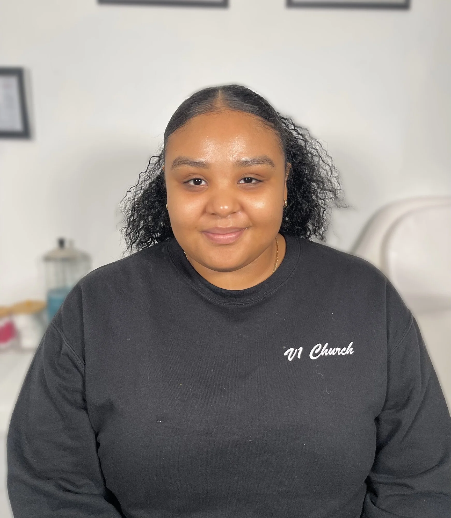 A young woman with curly black hair smiling, wearing a black sweatshirt with 'V1 Church' written on it, sitting in a room with a medical bed and blurred objects in the background.