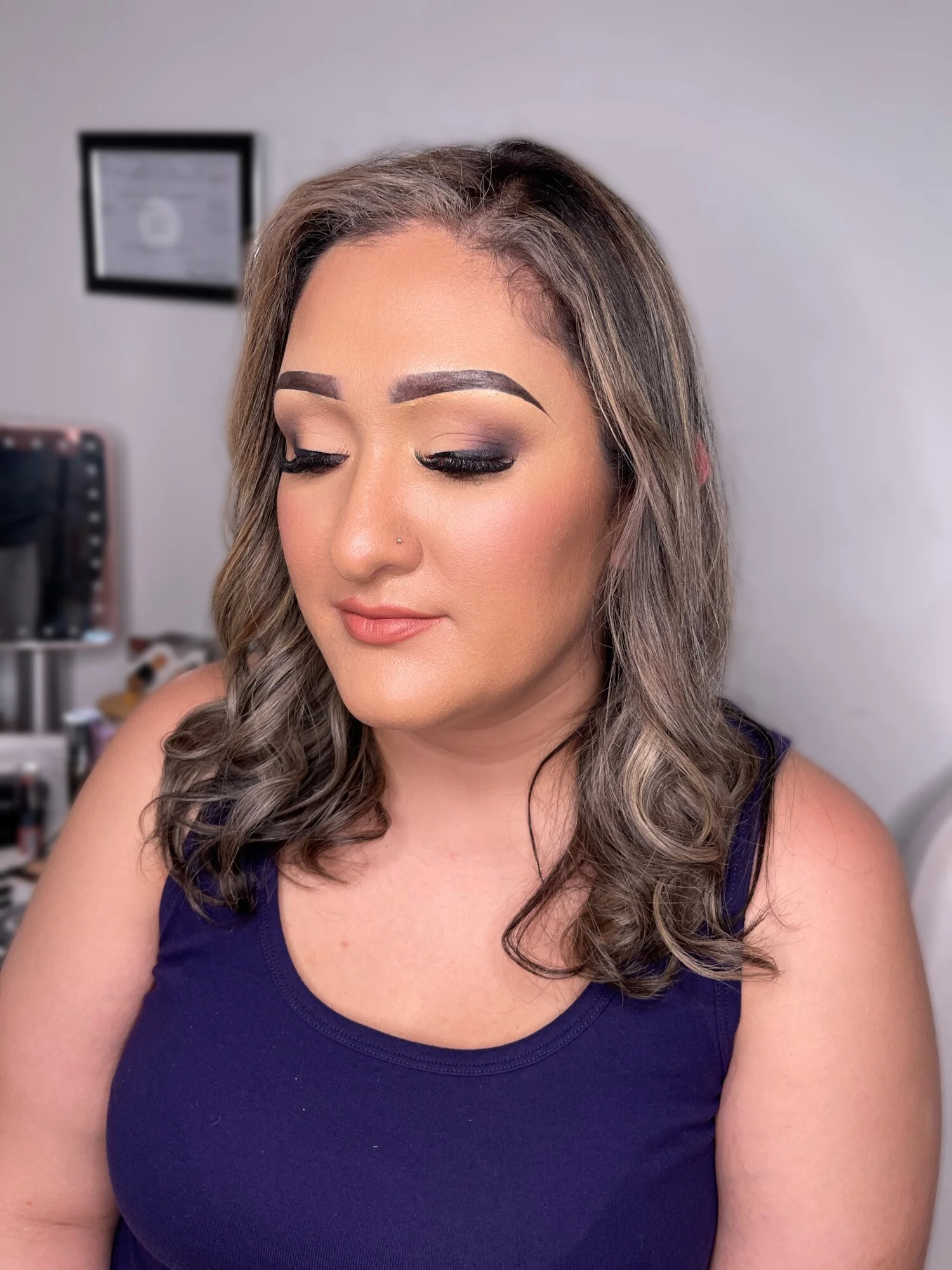 A woman with styled brown hair, makeup, and a nose piercing, wearing a blue sleeveless top, sits with her eyes closed in a room.