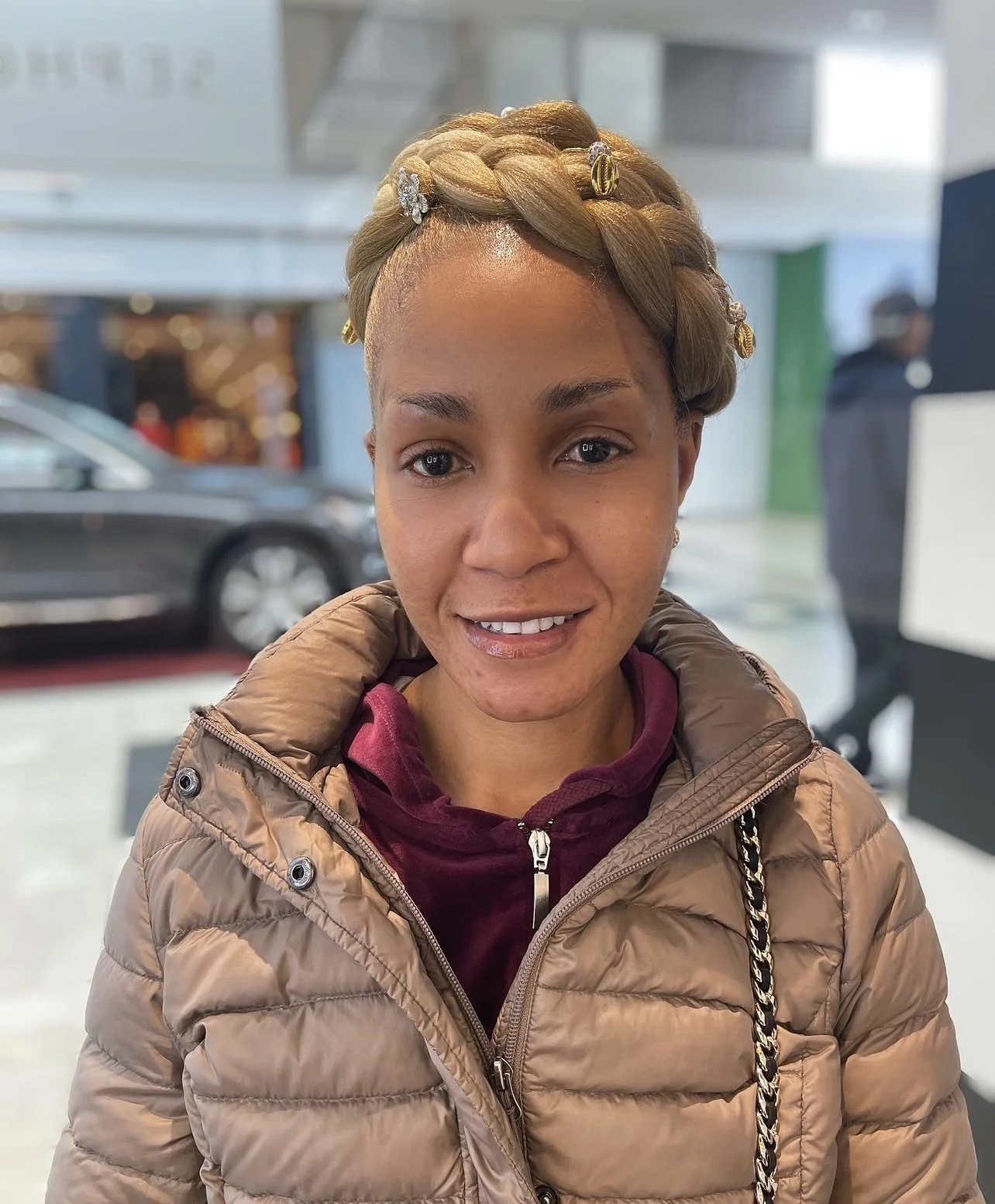 A woman with short, blonde hair styled in a braided crown adorned with shell and gold hair clips, wearing a beige puffer jacket over a burgundy hoodie, inside a brightly lit indoor space with cars and people in the background.