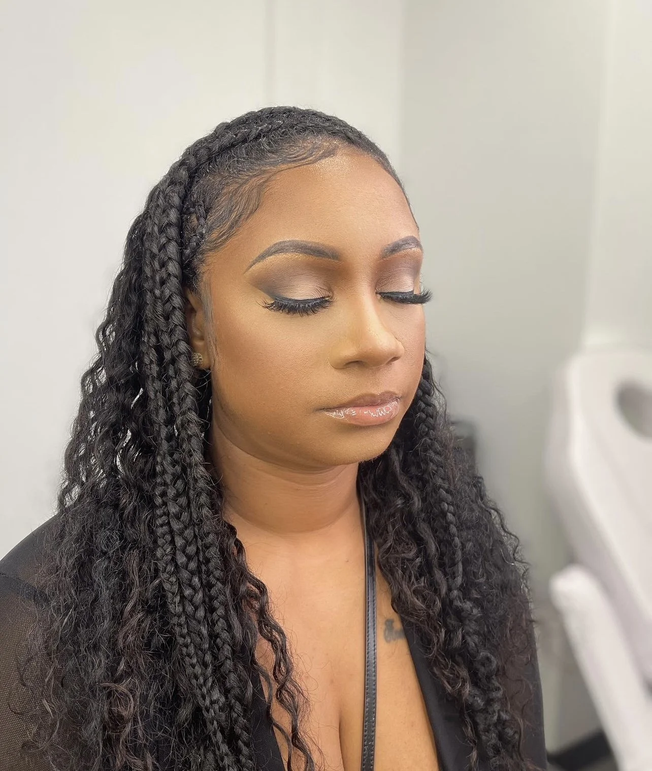 A woman with long, braided hair and makeup, sitting with her eyes closed in a neutral room with a white wall and medical equipment in the background.