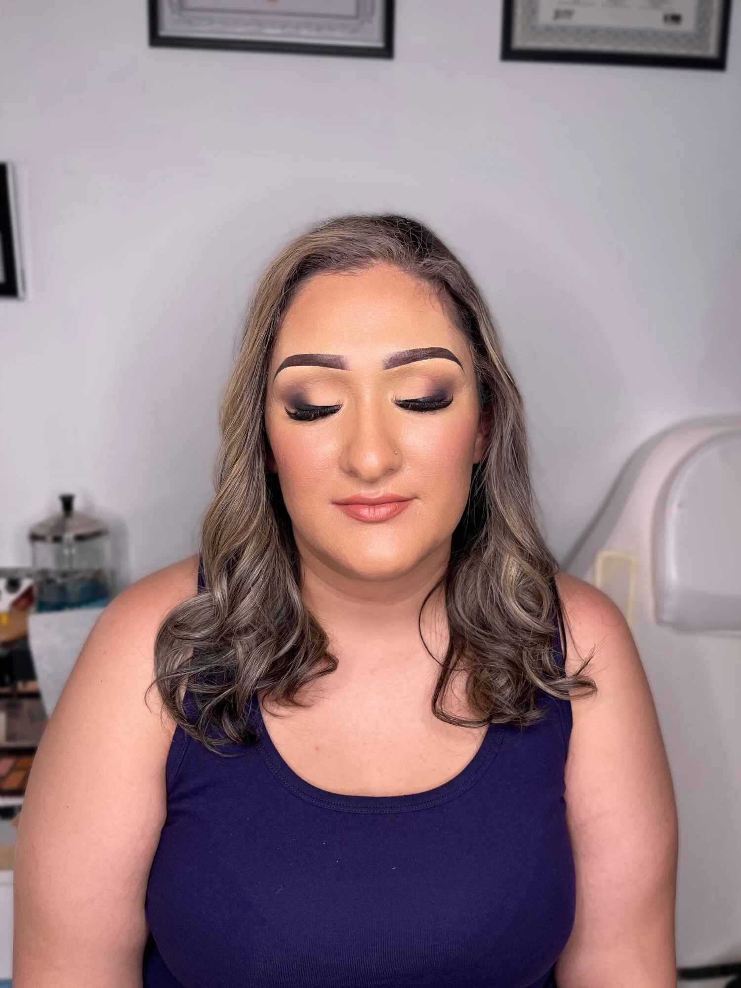 A woman with styled hair and makeup, wearing a navy blue tank top, has her eyes closed and is smiling softly in a makeup studio.