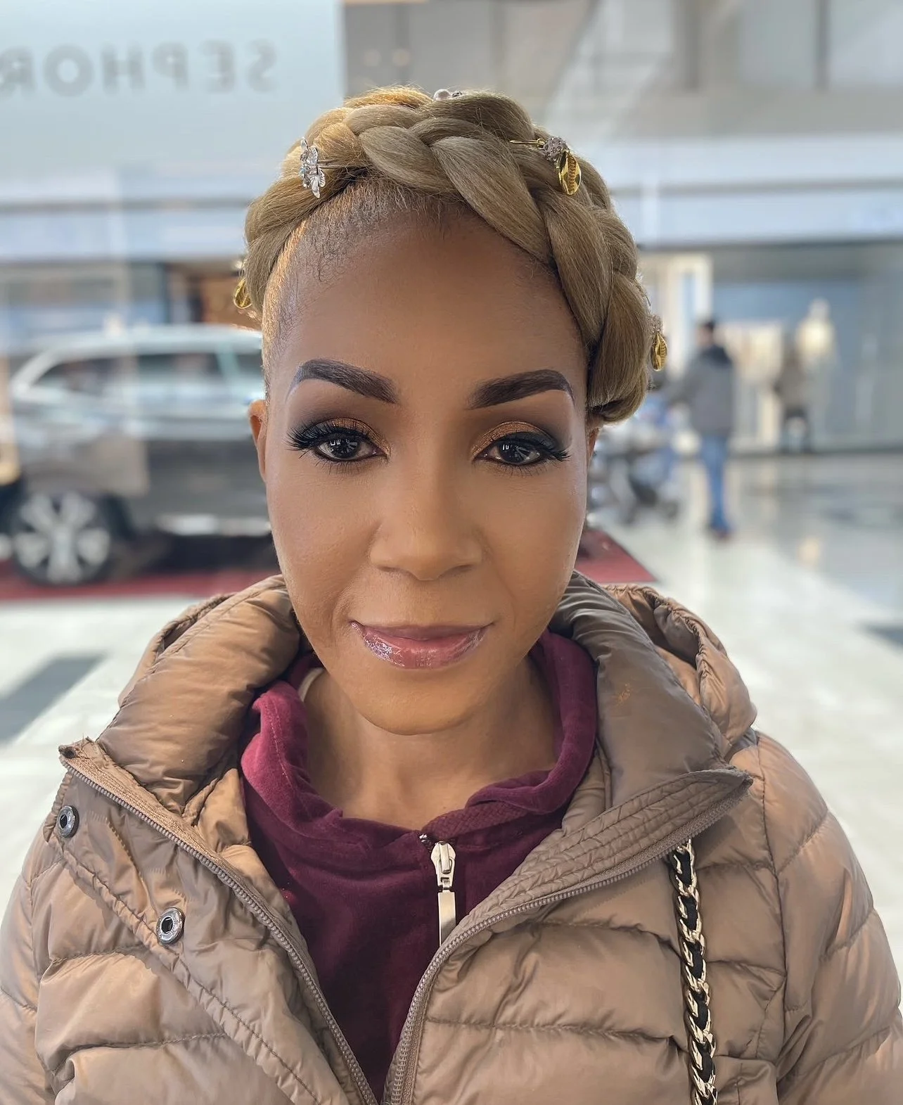 A woman with blonde braided hair styled with decorative hairpins sitting inside a building with large glass windows, wearing makeup, a beige puffer jacket, and a maroon hoodie.