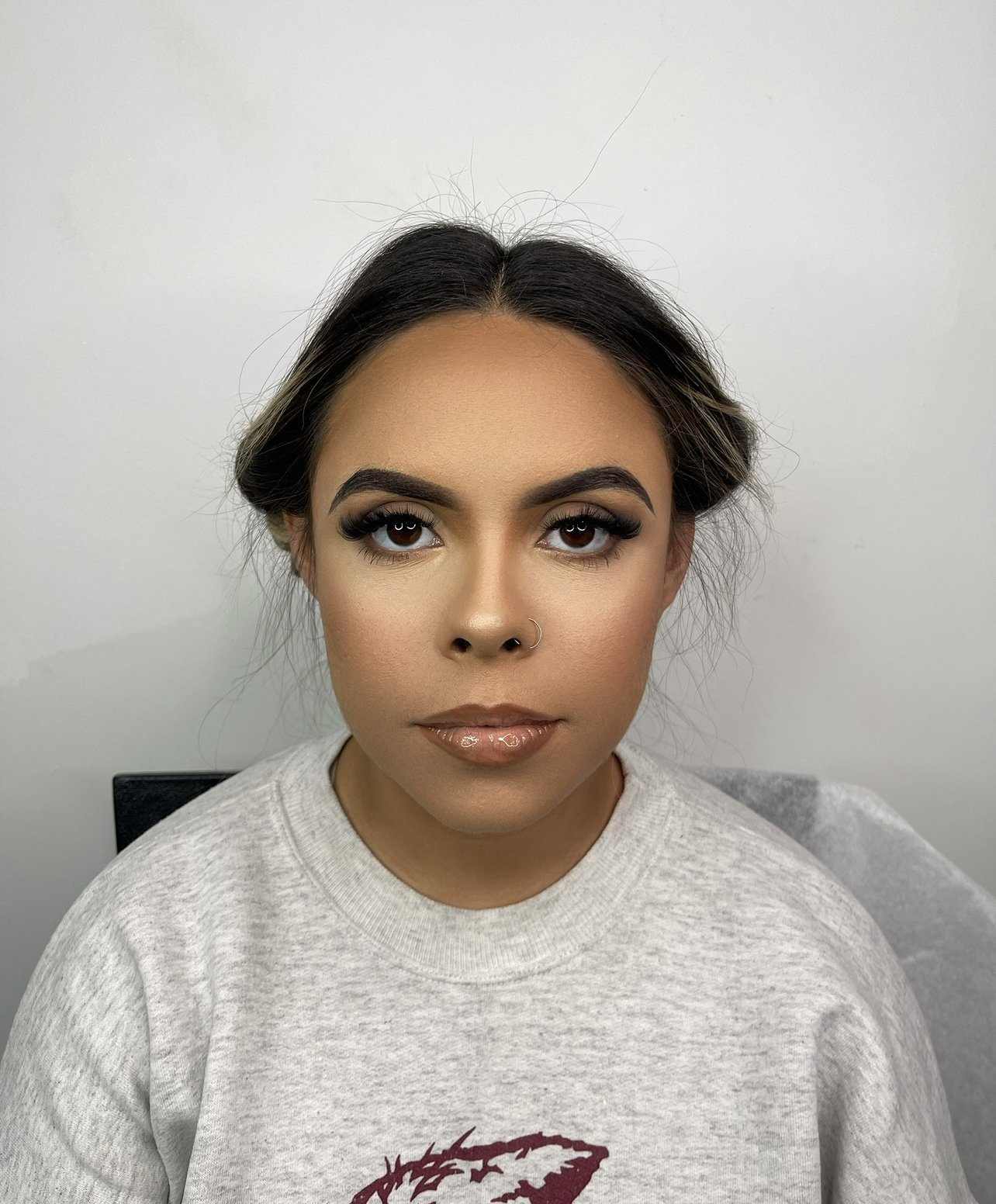 Close-up of a woman with makeup, dark hair, and a nose piercing, wearing a light gray sweatshirt with a red graphic, in front of a plain white wall.