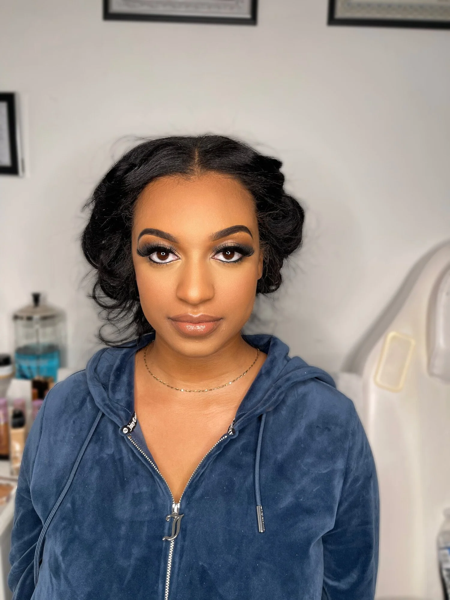 Close-up portrait of a woman with dark, wavy hair, wearing makeup and a navy blue zip-up hoodie, standing indoors in front of a white background with framed pictures and medical equipment.
