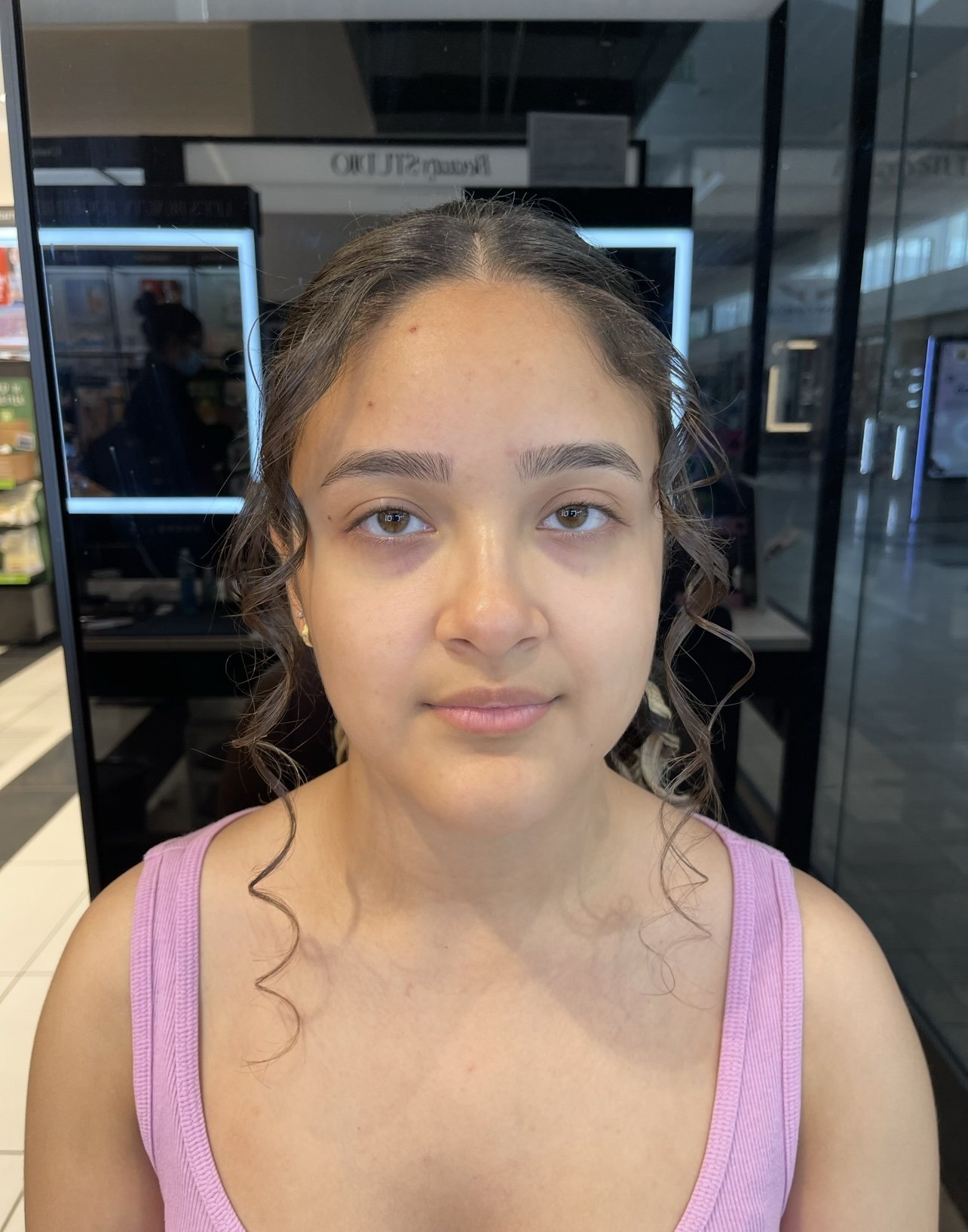 A young girl with light brown hair and hazel eyes, wearing a pink tank top, sitting in what appears to be a shopping mall or store, with electronic screens and glass doors in the background.