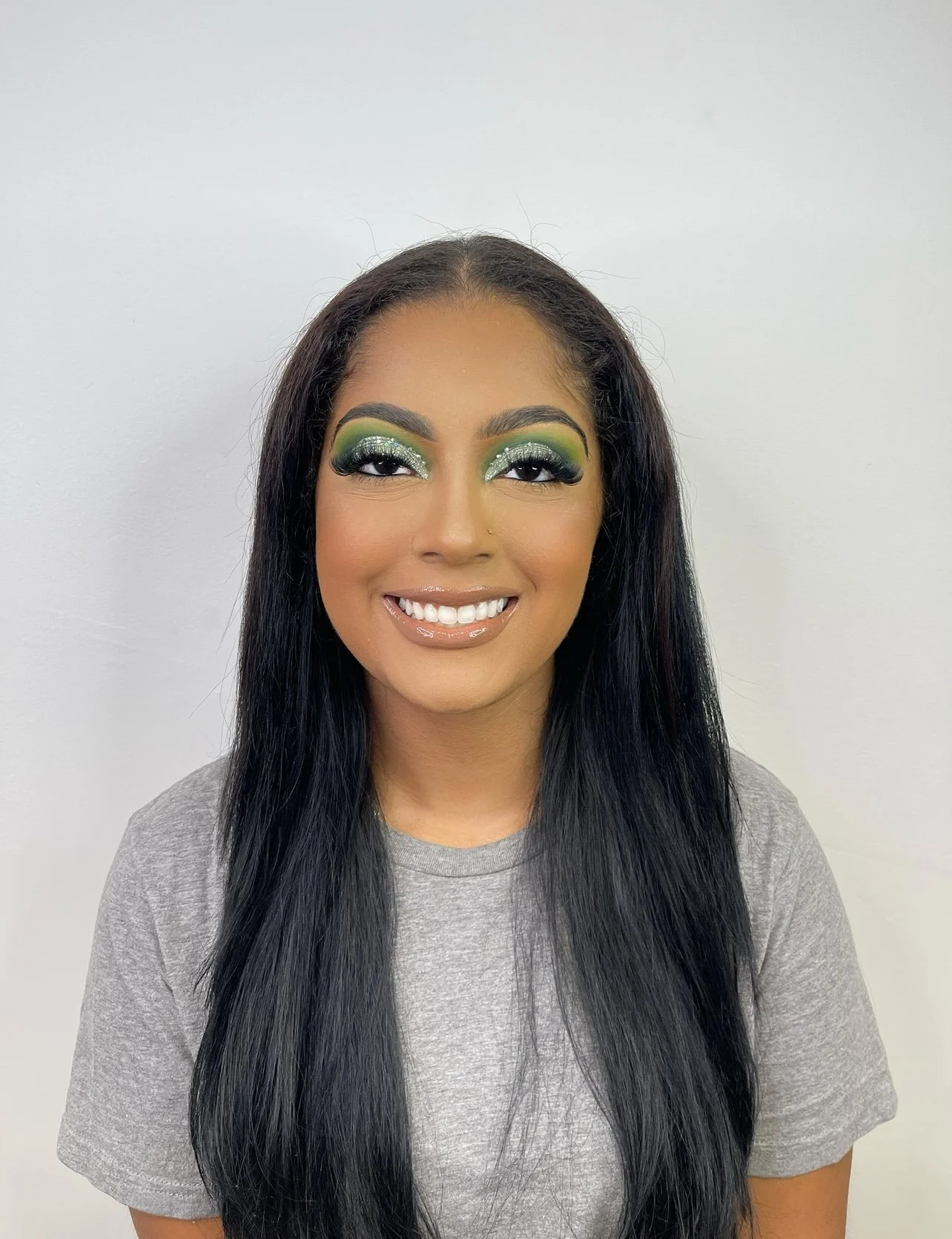 A woman with long black hair, smokey eye makeup with green and silver eyeshadow, and glossy lips, smiling at the camera against a plain white wall.