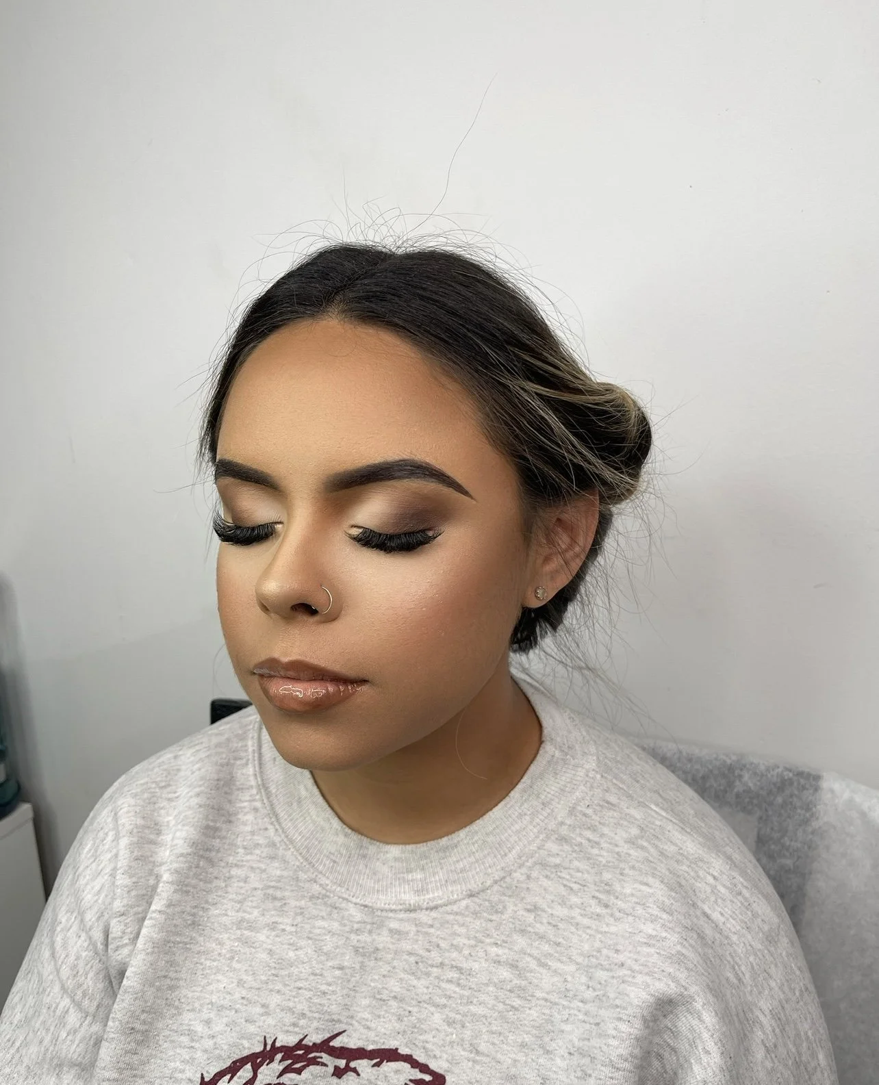 A woman with makeup, closed eyes showing eyeshadow and long eyelashes, wearing a gray sweatshirt with a graphic, small earrings, and a nose ring, sitting against a white wall.