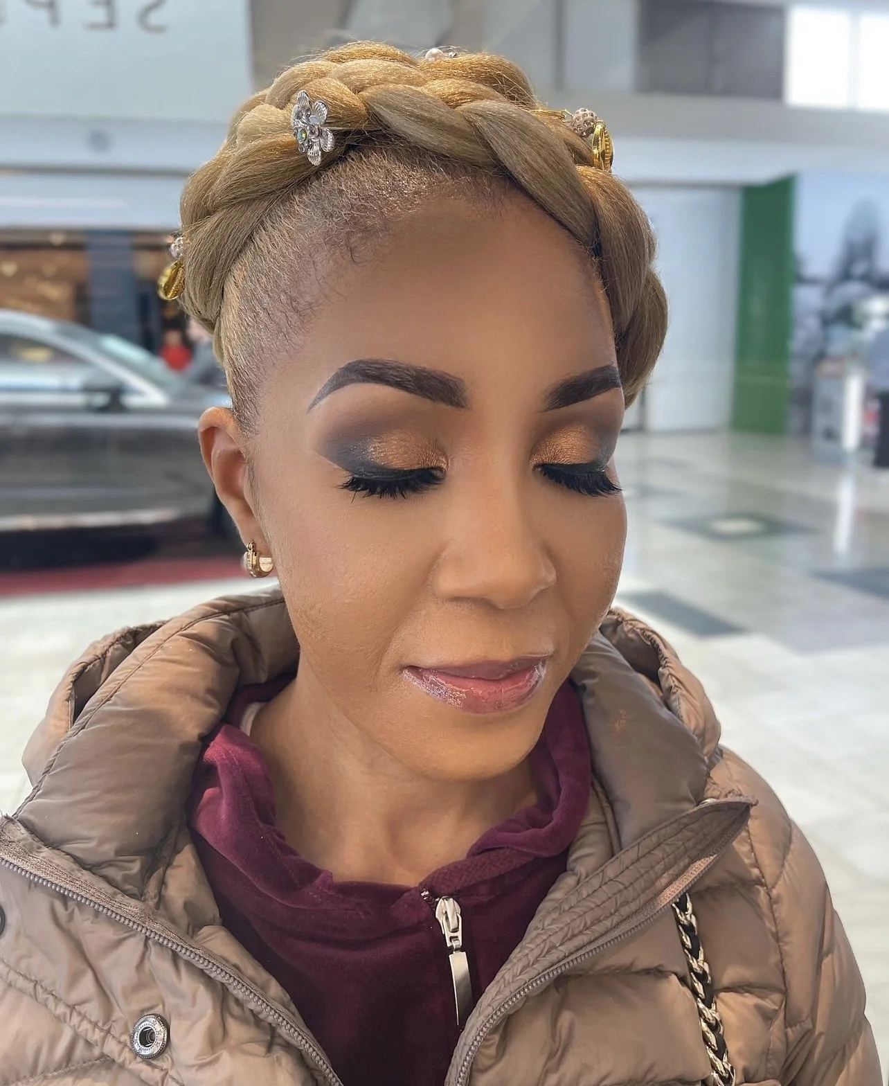 A woman with styled blonde hair wearing decorative pins, closed eyes showing gold and dark eyeshadow, with glossy lips, dressed in a brown puffer jacket and a maroon hoodie.