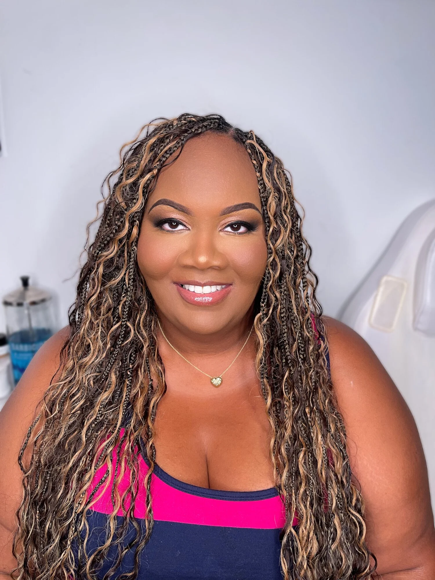 A woman with long, curly hair wearing a pink and navy sleeveless top, gold necklace, and smiling in a professional setting.