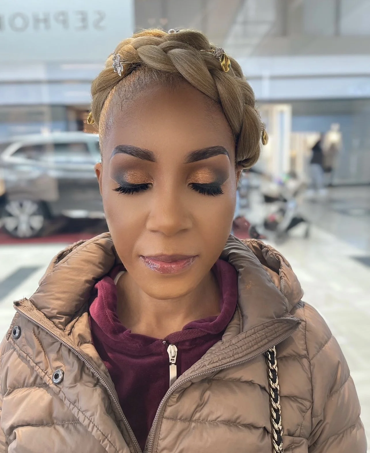 A woman with closed eyes, makeup, and braided hair decorated with butterfly and gold hairpins, wearing a brown jacket and purple hoodie inside a modern building with cars and people outside.