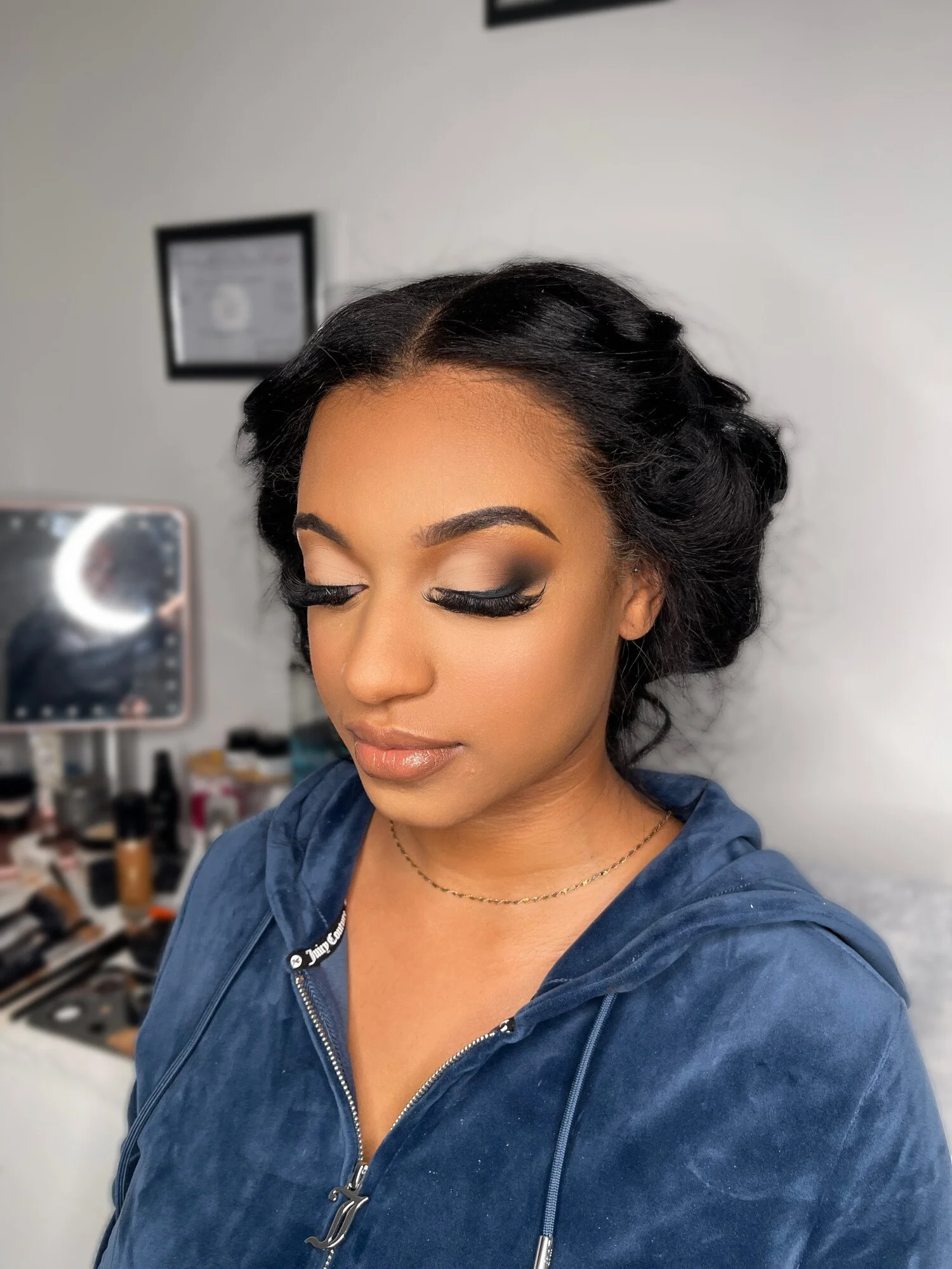 A woman with dark hair styled in waves, wearing makeup with dramatic eye makeup, false eyelashes, and nude lip gloss. She is wearing a navy blue zip-up hoodie and a delicate gold necklace. The background shows a blurred mirror, makeup products, and a