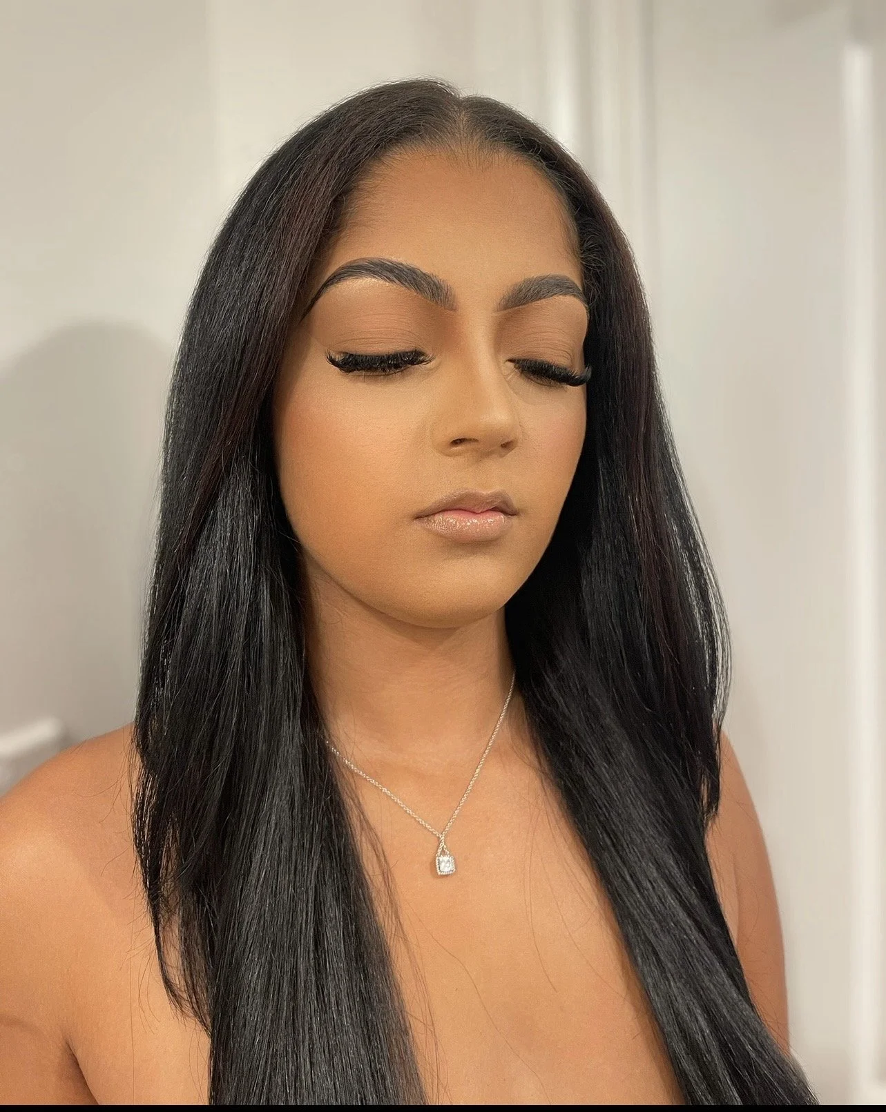 A young woman with long black hair, closed eyes, makeup, and a necklace with a pendant, standing against a neutral background.