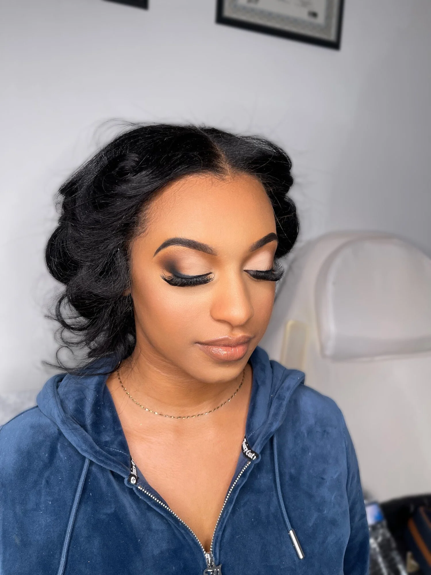 Close-up of a woman with makeup, black hair, and a blue zip-up hoodie, with a blurred background of a white wall and partially visible framed pictures.