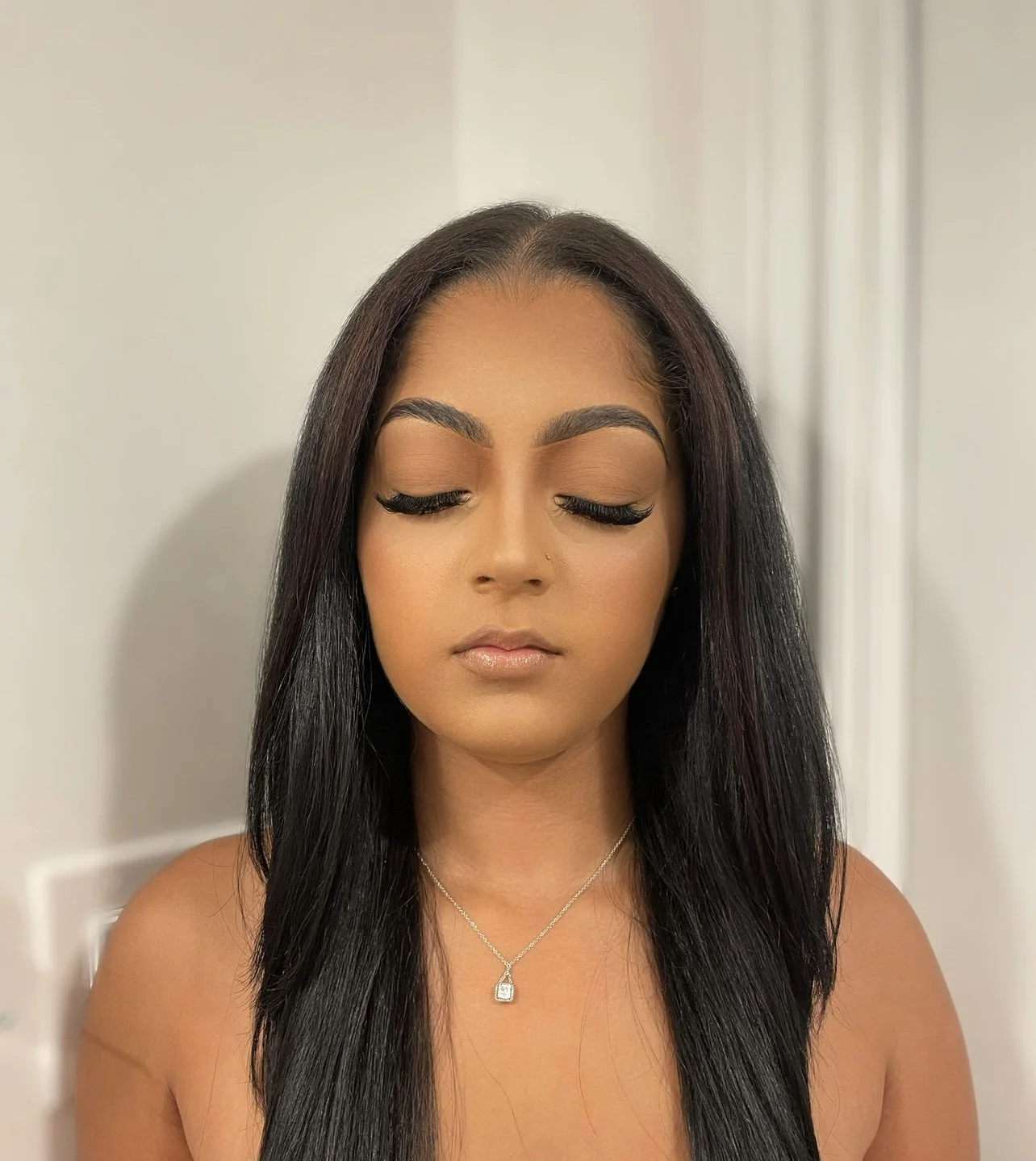 A young woman with long black hair and medium skin tone, wearing makeup with prominent eyelashes, a necklace with a pendant, and a neutral expression, standing indoors against a plain light-colored wall.