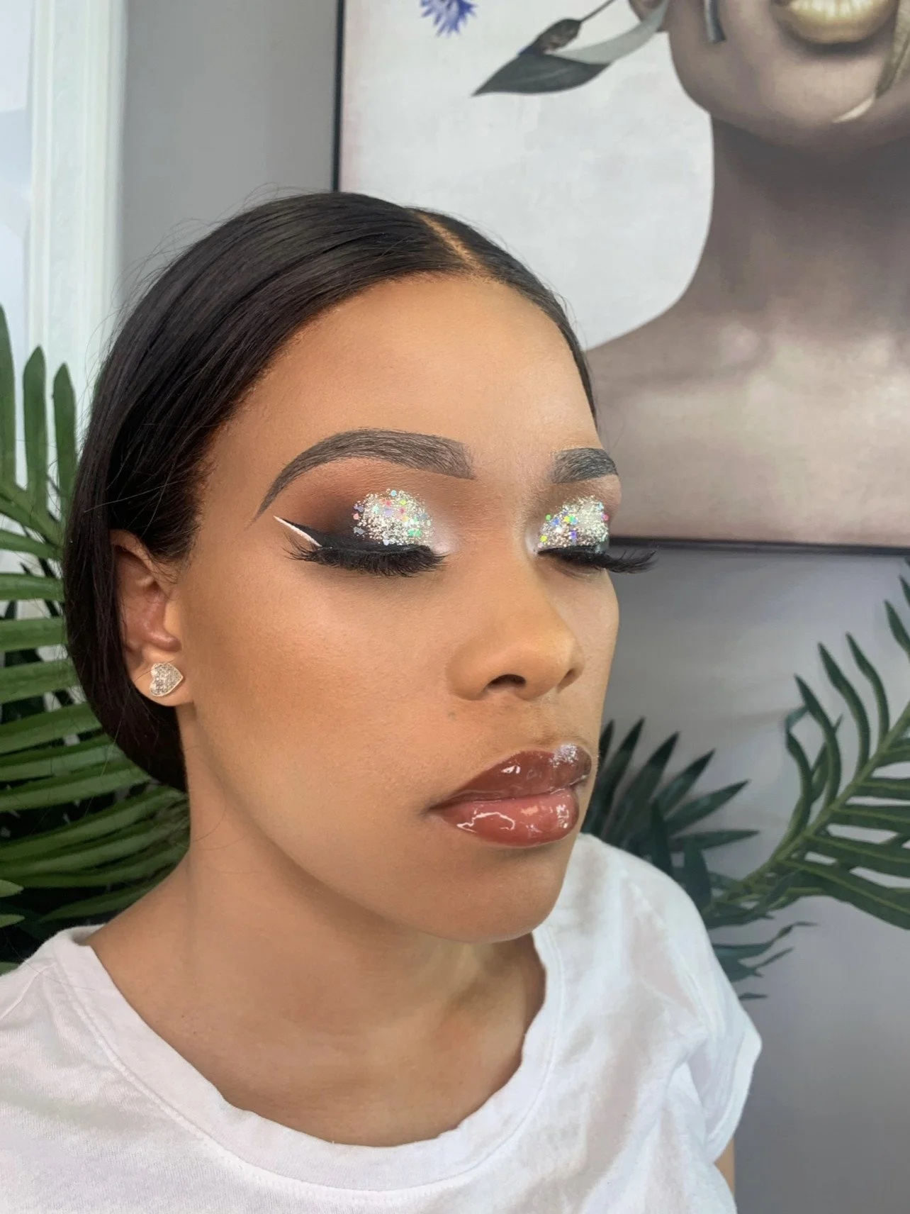 Close-up of a woman with makeup, glitter eyeshadow, and glossy lips, with a painting of a woman with jewelry and makeup in the background.