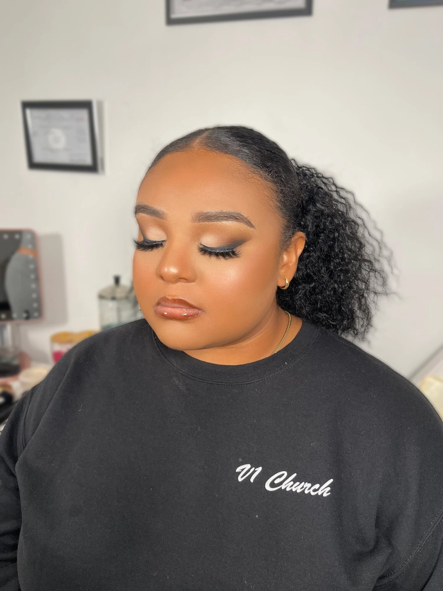A woman with styled curly hair, wearing makeup, earrings, and a black sweatshirt with white text, standing in a room with framed pictures on the wall and makeup items in the background.