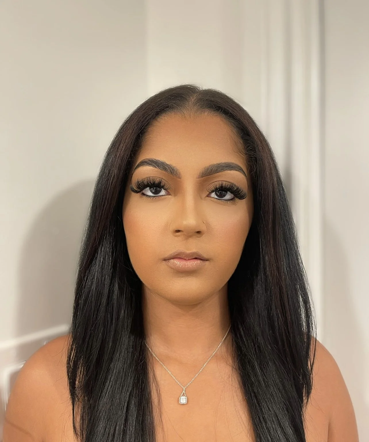 A woman with long black hair, dark eye makeup, and a necklace, looking directly at the camera against a plain light-colored background.