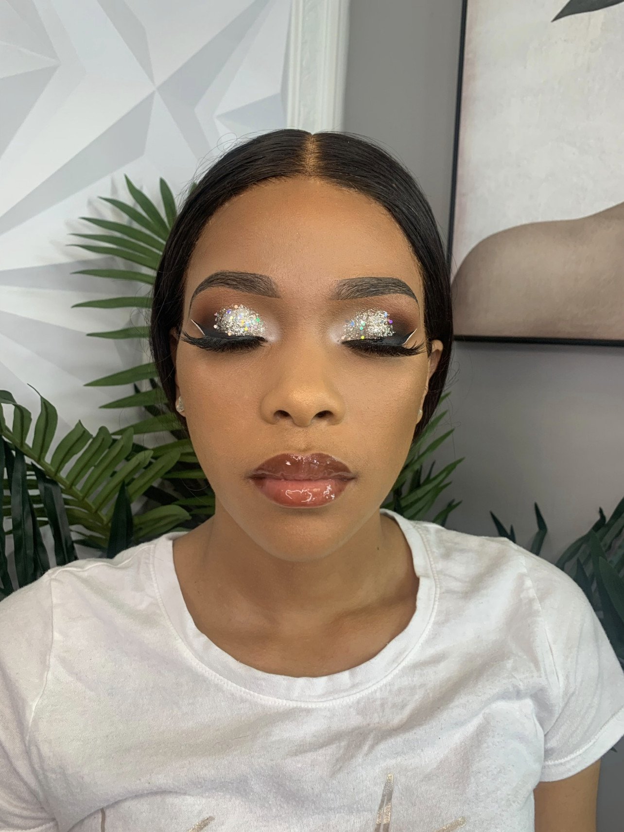 Close-up of a woman with makeup, including glittery eyeshadow, long eyelashes, and shiny lips, with plants and artwork in the background.