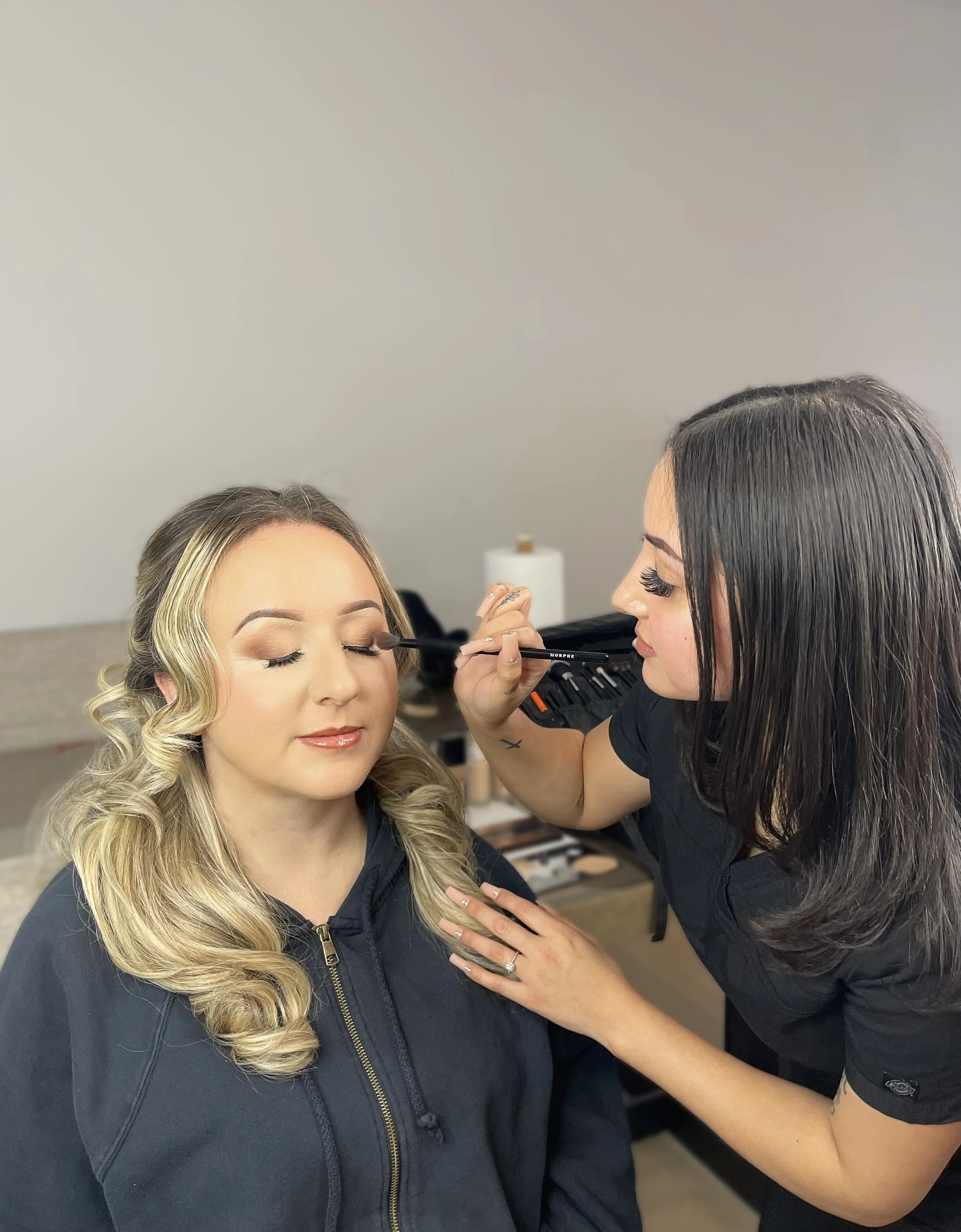 A woman with long, styled blonde hair is getting her makeup done by a makeup artist. The woman has her eyes closed and appears relaxed, while the makeup artist is applying makeup near her eye.