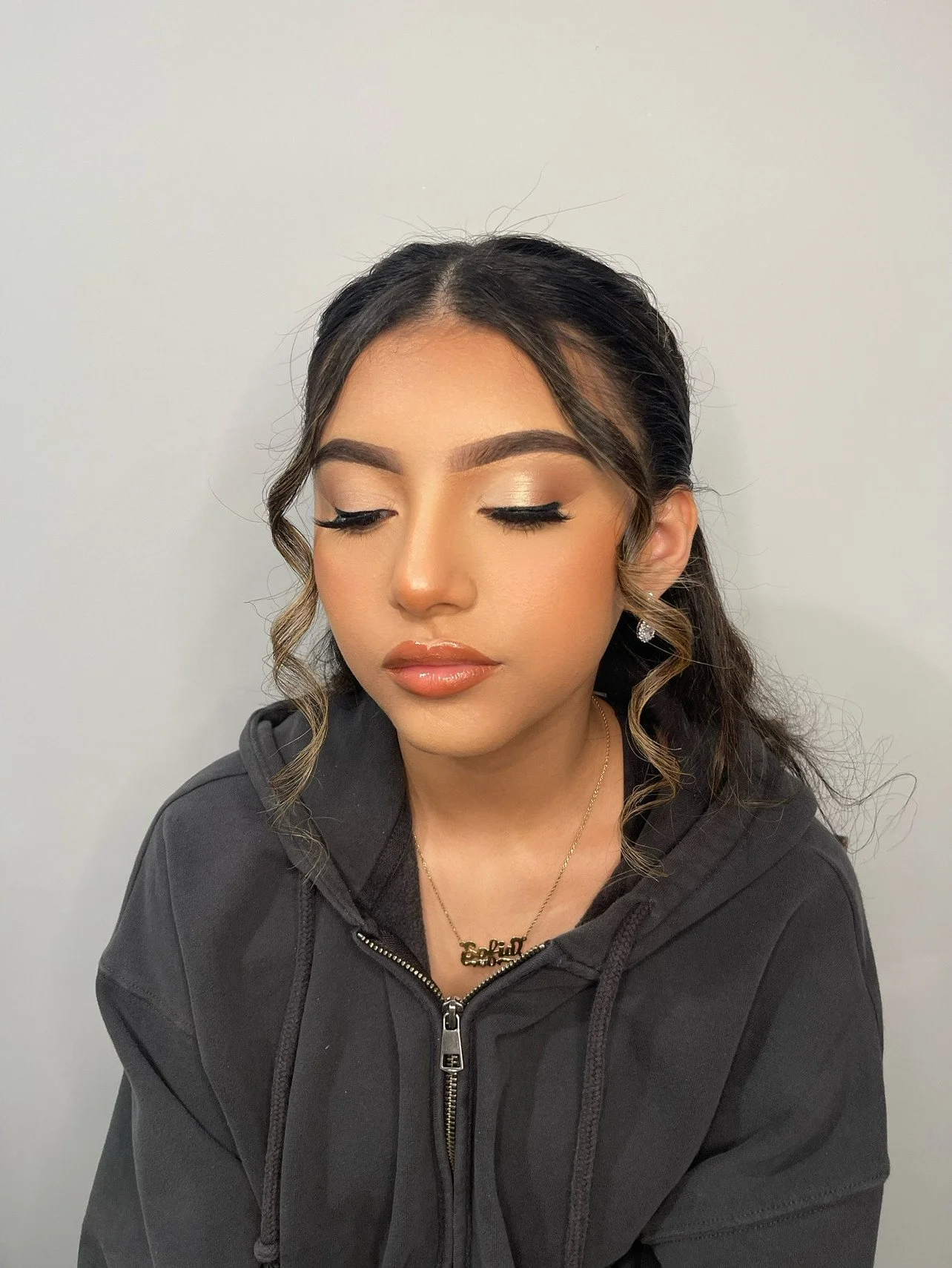 Young woman with styled hair, makeup, and jewelry, wearing a black hoodie, with eyes closed against a plain white background.