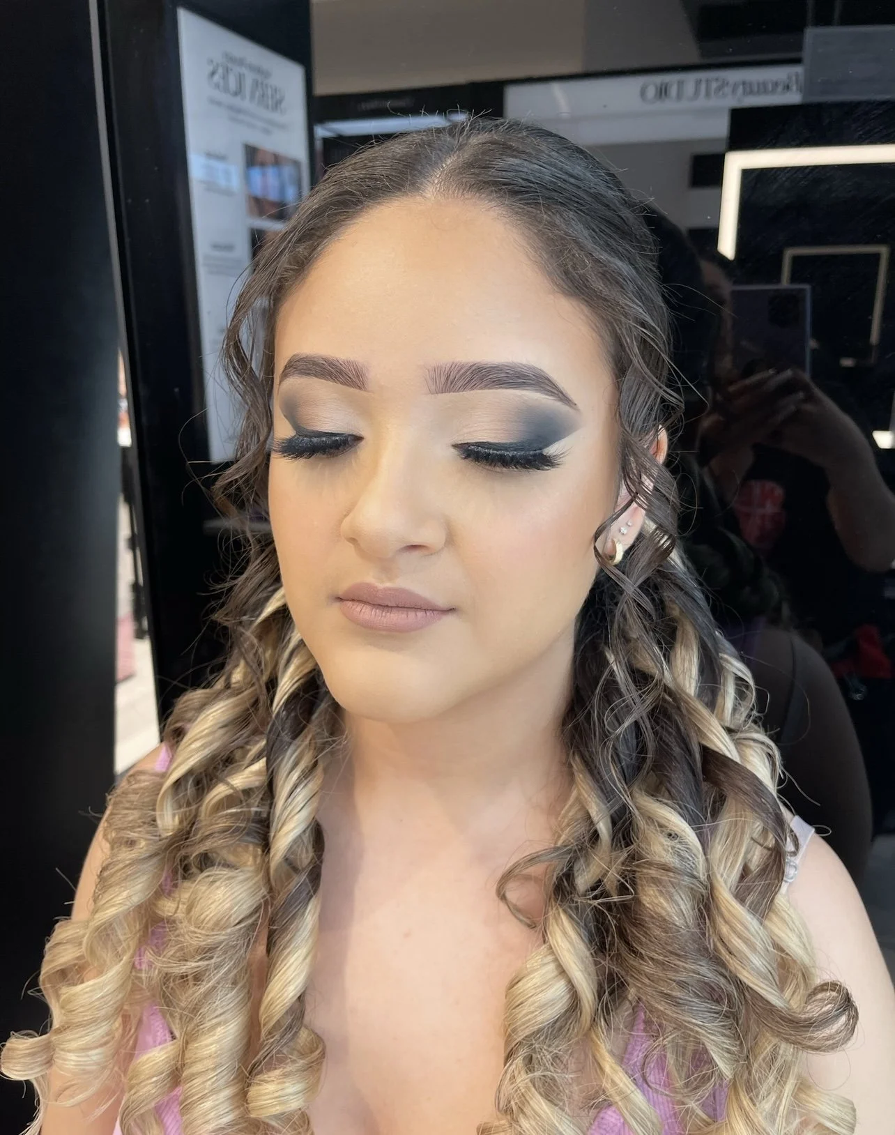 Close-up of a woman with long, curly blonde and brown hair, wearing makeup with smoky eyeshadow, eyeliner, long lashes, and nude lipstick, in a makeup studio.