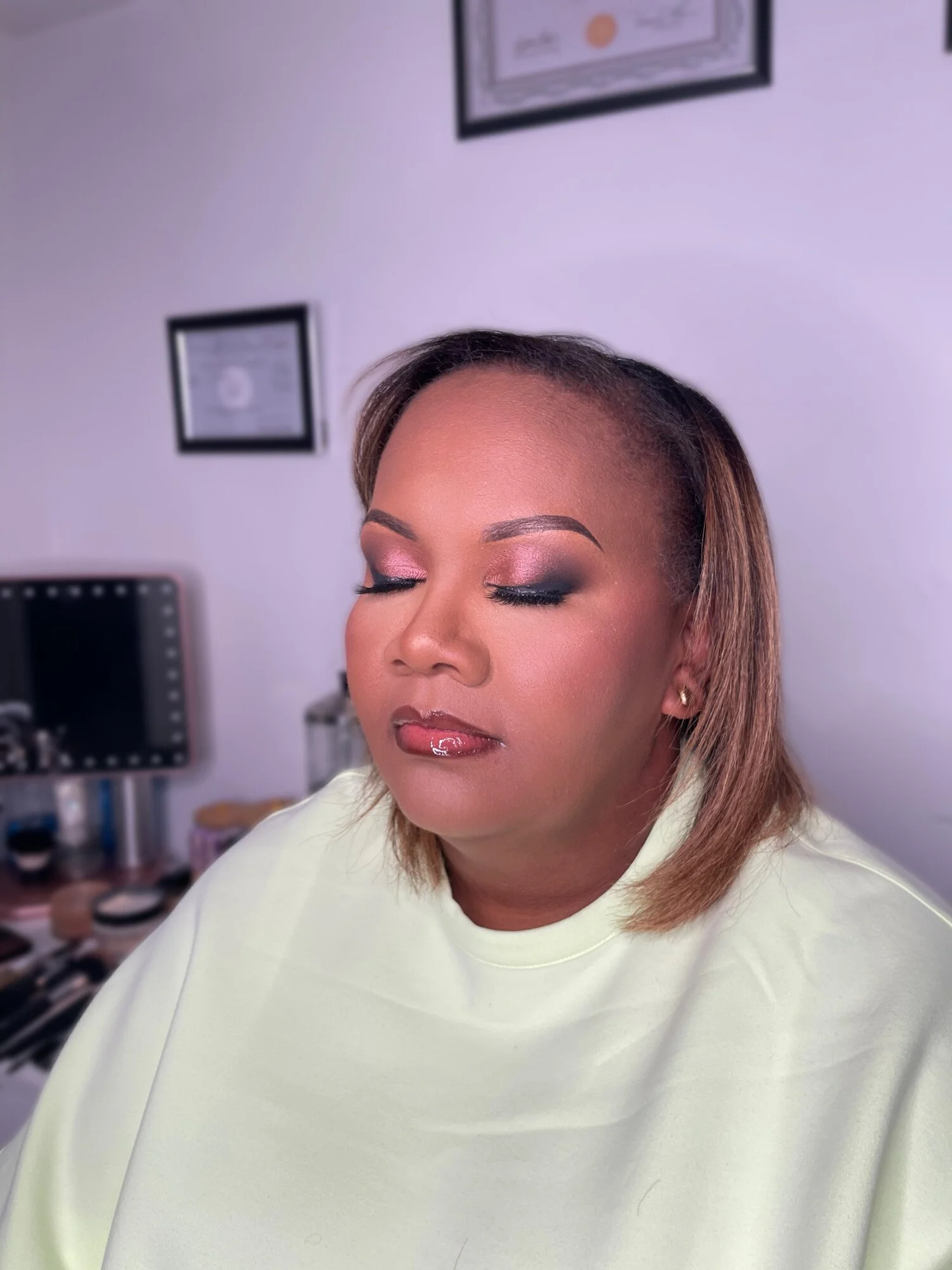 A woman with makeup and closed eyes is in a makeup studio, wearing a light-colored shirt.