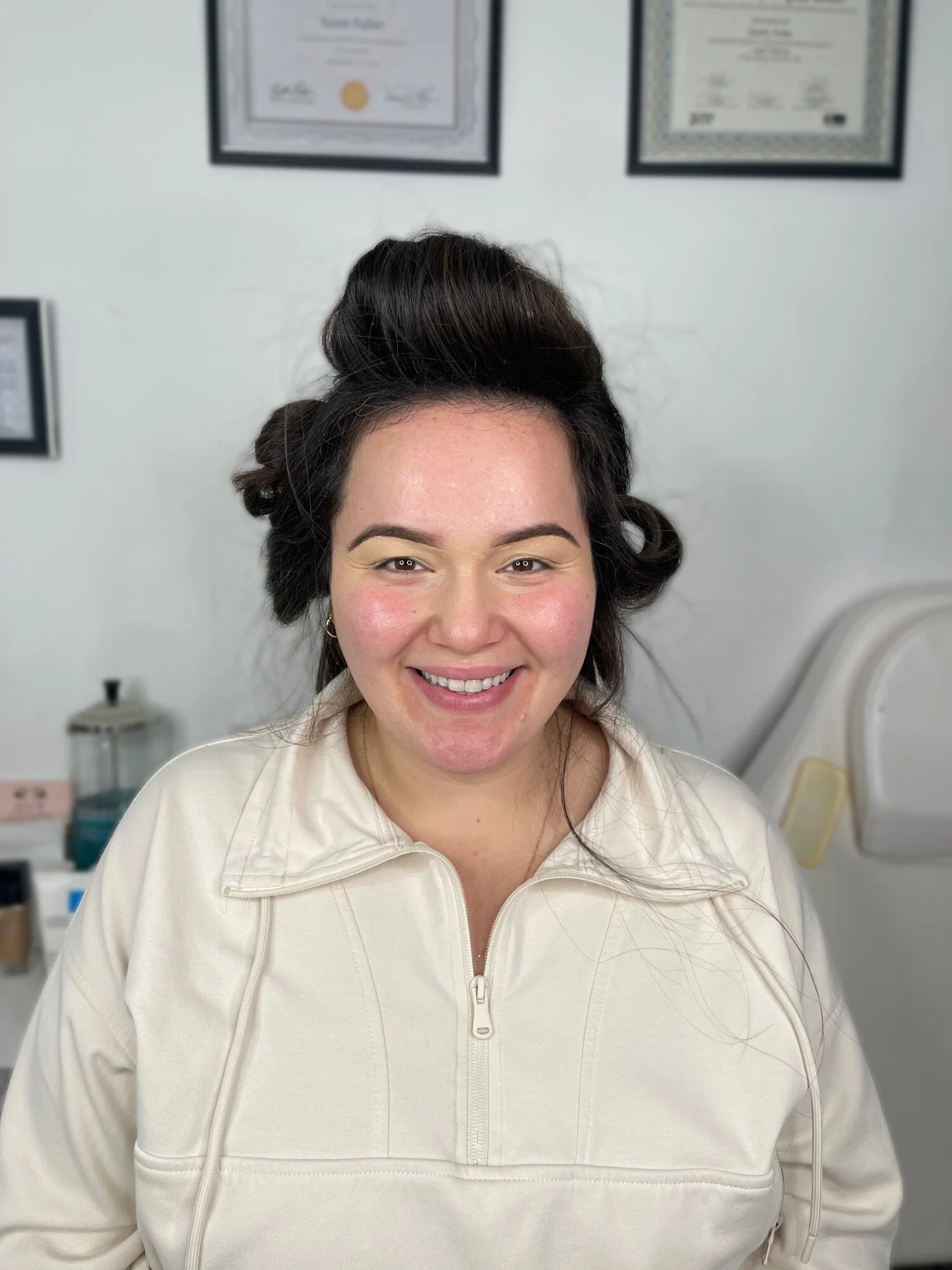 A woman with dark hair styled in vintage curls smiling at the camera, wearing a cream-colored zip-up jacket, in a room with framed certificates on the wall.
